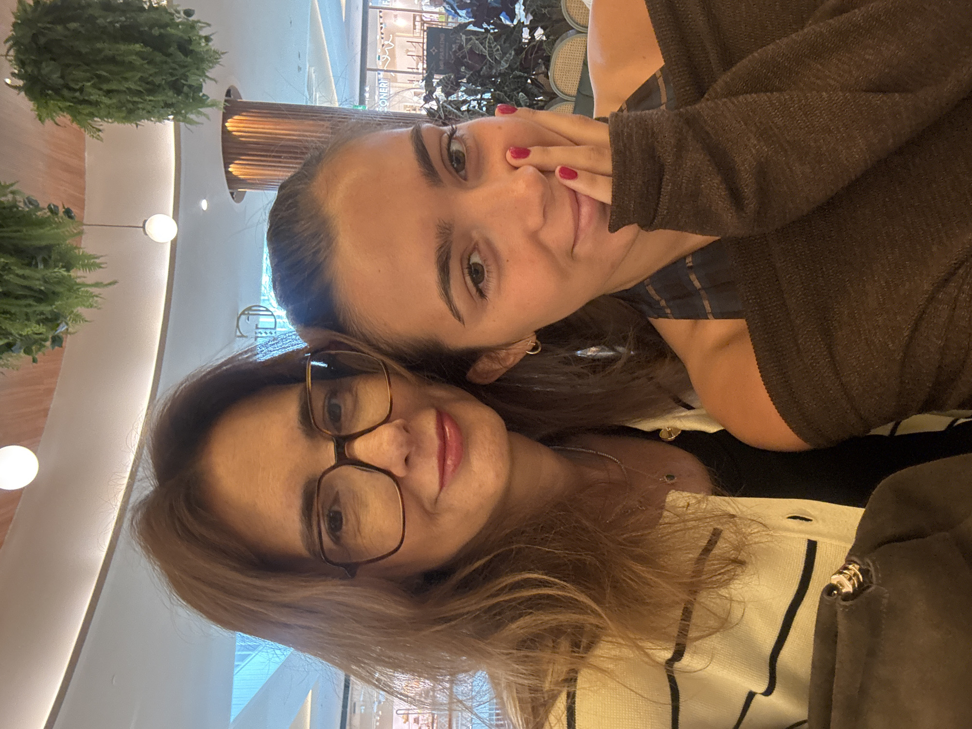 Two women are posing closely together for a selfie indoors. The woman in the foreground has shoulder-length brown hair, eyeglasses, and is wearing a light-colored, striped sweater. Behind her, the other woman has her hair pulled back, is wearing a brown sweater, and has red nail polish. The background includes indoor foliage, decorative lighting, and reflections of storefronts. This location is likely a shopping center in Senhora da Hora, Portugal.