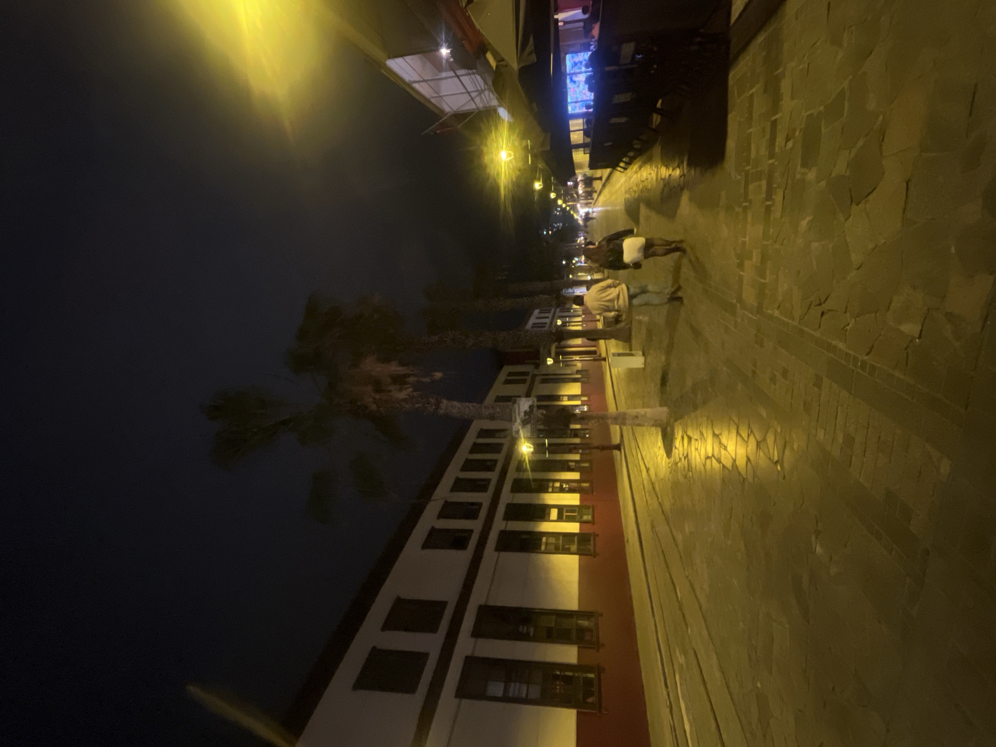 The image depicts a street scene at night in the city of Iquique, Chile. The street is paved with irregular flagstones that are wet and reflect the yellow light from street lamps. On the left side of the street, there is a long, white building with a red lower facade and dark rectangular windows. Palm trees line the street, with their silhouettes visible against the dark night sky. Two individuals, a man and a woman, are walking away from the camera down the center of the street. The man is wearing a light-colored jacket and jeans, while the woman is wearing a dark top and a white skirt. To the right of the street, there are outdoor seating areas for restaurants, with tables and umbrellas visible. The overall atmosphere is calm and somewhat atmospheric due to the night lighting and the wet pavement.