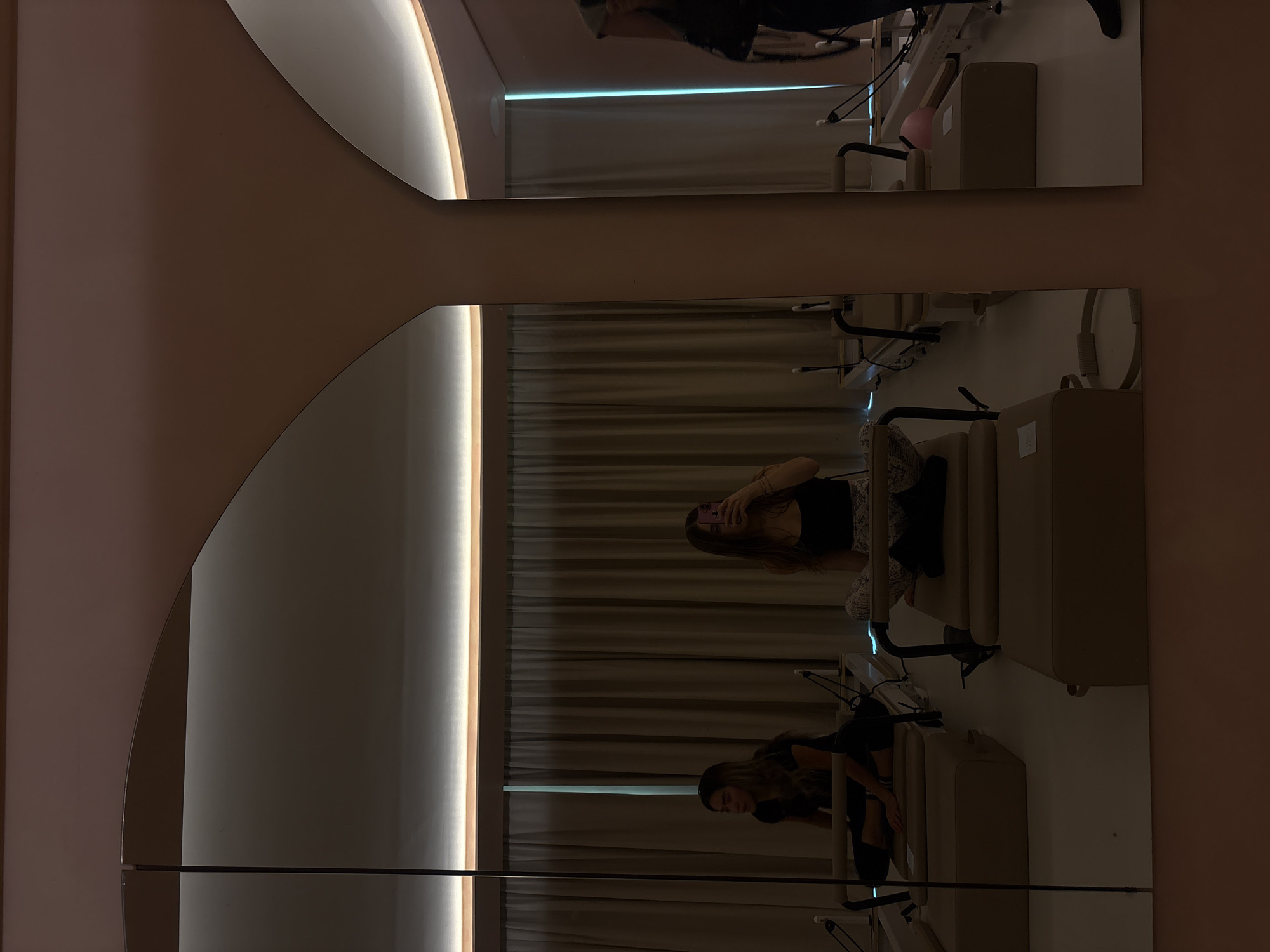 A woman is reflected in two mirrors in a room. The mirrors are affixed to a wall. The woman is wearing a black tank top, a patterned pair of pants, and black shoes. She is sitting on a padded reformer machine and holding a phone in her hands. The reformer is beige in color. The background behind the woman is a beige curtain. The room is located in Mexico City, Mexico.