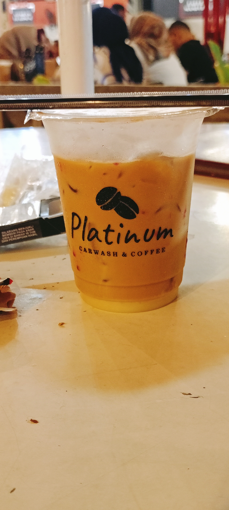The image captures a plastic cup of iced coffee from "Platinum Carwash & Coffee," as indicated by the logo on the cup. The coffee has a layer of milk at the bottom and what appears to be ice and possibly coffee grounds floating within. The cup is sealed with a plastic lid. To the left of the coffee is a packet of cigarettes or similar items. In the background, partially obscured, are several people seated at tables, possibly at the café. The location is Lhokseumawe, Indonesia, based on the local business and the Indonesian warning label visible on the packet near the coffee. The environment appears to be a casual café setting, possibly part of the Platinum carwash and coffee shop.