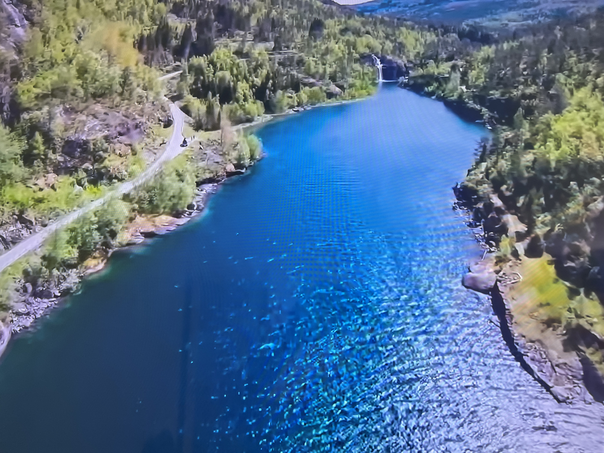 Aerial shot of a fjord or long lake surrounded by forested hills, likely in Norway. The water is a vibrant blue with clear visibility and ripple patterns. A road follows the shoreline on the left. A waterfall is visible in the distance at the far end of the lake. The image does not appear to be in Basingstoke and Deane, United Kingdom.