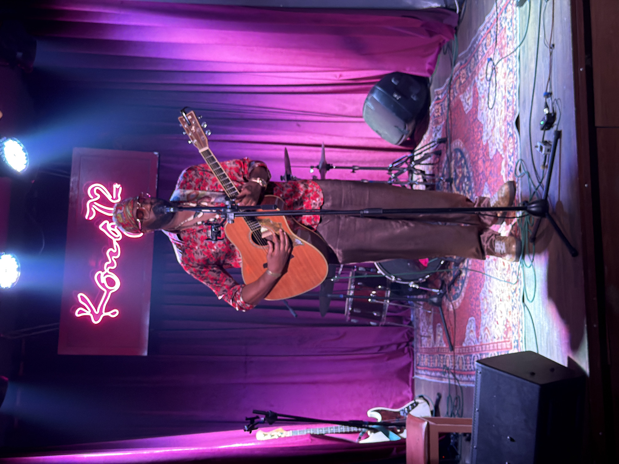 This indoor photograph captures a male musician performing live on stage, likely at a music venue in Đà Nẵng, Vietnam, during the evening or night. The image is originally oriented 90 degrees clockwise.
The central figure is a man playing an acoustic guitar, positioned towards the right side of the frame. He wears a patterned red and white long-sleeved shirt, dark trousers, a patterned head covering, and sunglasses. A microphone on a stand is placed directly in front of him, suggesting he is also singing.
The stage is set against a backdrop of purple drapes or curtains, with the floor covered by a decorative red patterned rug. The ambiance is created by dramatic stage lighting, featuring prominent spotlights emitting blue and purple hues from the left side of the stage.
A bright pink neon sign spelling out "Kongg" is visible on the left side of the stage, adding to the venue's character. Other musical equipment includes a partially visible drum kit behind the performer, a bass guitar resting on a stand near the front left of the stage, and a speaker monitor on the far right. The musician appears focused on his performance, contributing to an intimate and engaging atmosphere.
