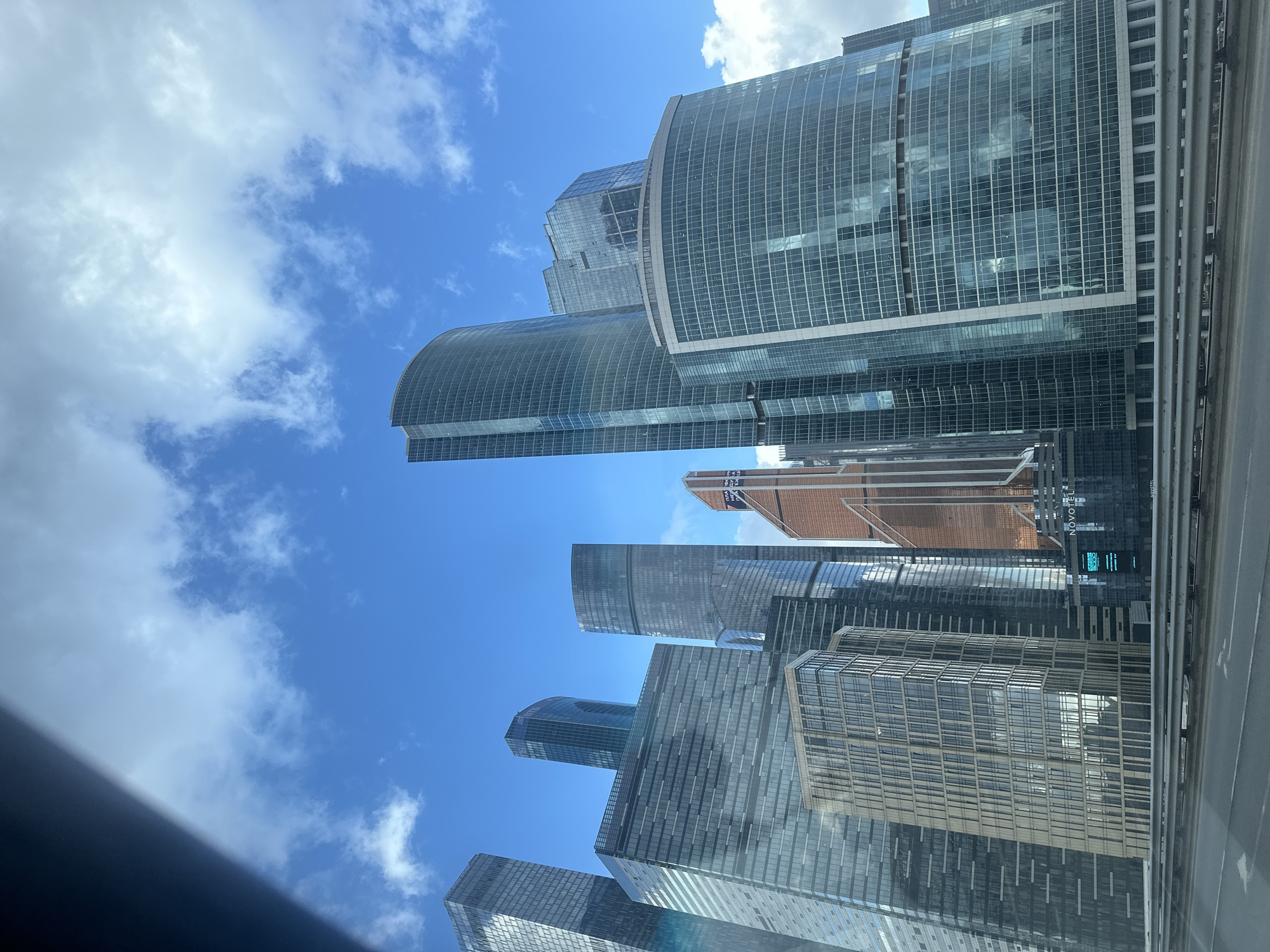 The image shows a view of the Moscow International Business Center, also known as Moscow City, in Moscow, Russia. The scene is taken from a low angle, looking upwards at the cluster of modern skyscrapers. The buildings are characterized by their glass facades, varying heights, and diverse architectural designs. Some have rounded or curved shapes, while others are angular. The sky is blue with scattered white clouds. The structures appear to be situated adjacent to a road or elevated platform. A partial frame of the vehicle from which the picture may be taken is visible at the bottom.