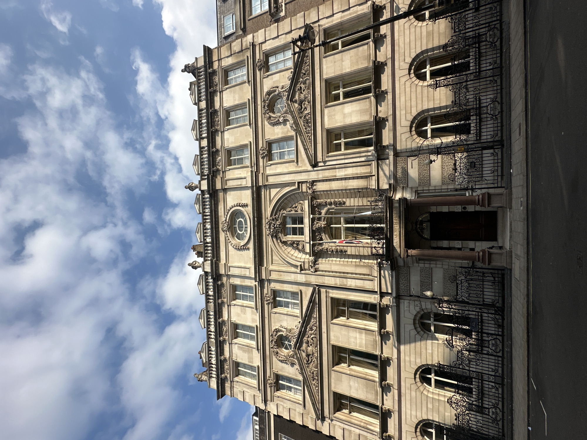 The image shows the exterior of a grand, multi-story stone building in London, United Kingdom, viewed from a low angle looking up towards the sky. The building's facade is ornate, featuring classical architectural elements such as columns, pediments, intricate carvings, and decorative sculptures along the roofline. Several windows with white frames are visible across the different levels. A balcony with wrought-iron railings is positioned above the entrance, adorned with a Union Jack flag. The building appears to be an older, established structure, possibly a historic government building or a large office building.
In front of the building, there is a dark wrought-iron fence with decorative scrollwork. A paved sidewalk runs along the front of the fence, leading to a street with asphalt surfacing. A lamppost stands to the right of the building, casting a shadow. The sky is a bright blue with scattered white clouds, suggesting a clear day with sunlight. The architecture and the presence of the Union Jack flag indicate a location within the United Kingdom. The signage on the building, though partially obscured, includes "DARTMOUTH HOUSE" and a street number "37", suggesting a specific address.