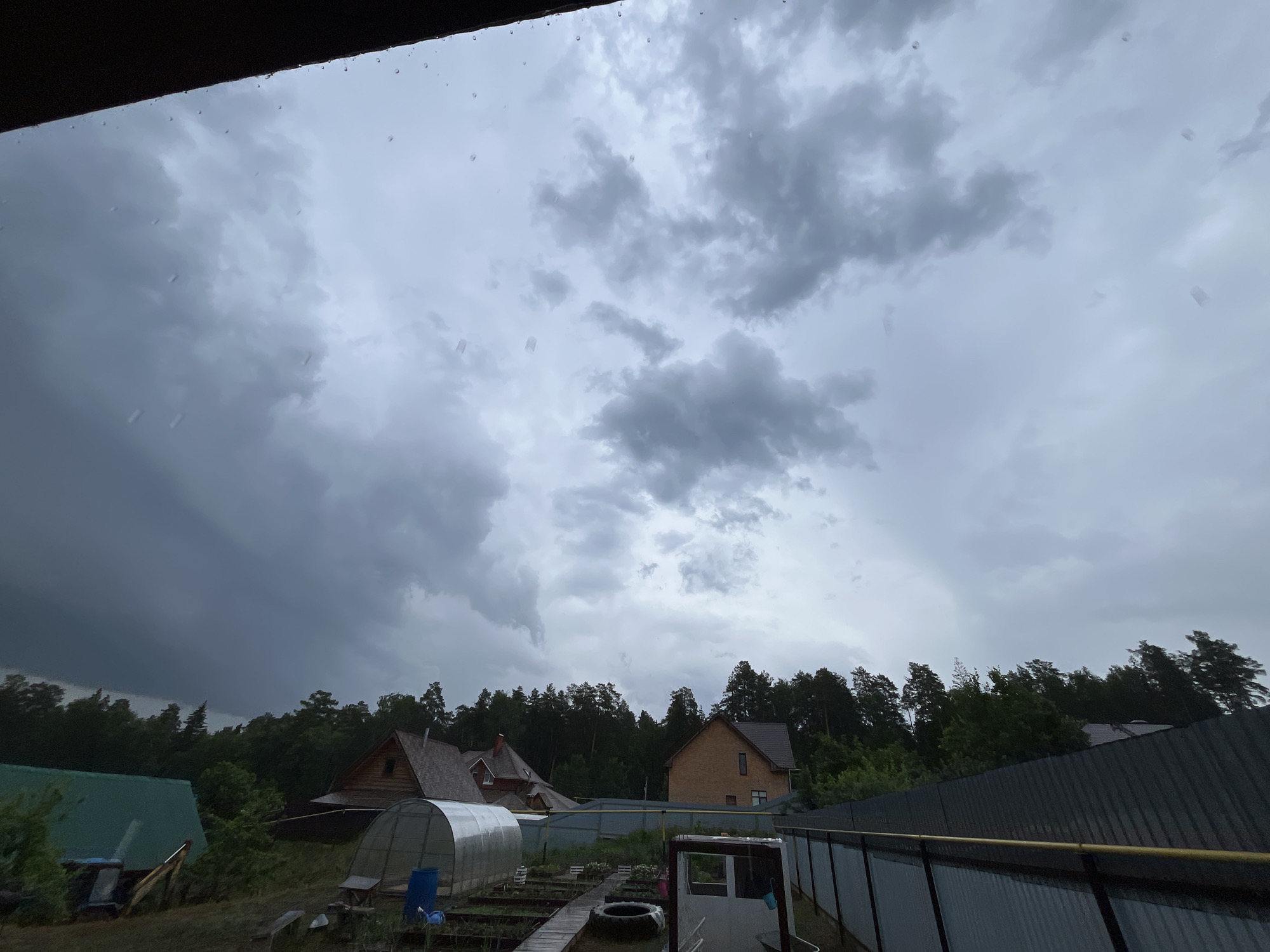The image shows an overcast sky with heavy, gray clouds, likely indicating rain. Raindrops are visible near the top of the frame, suggesting the picture was taken during a downpour. Below the sky, there's a view of a rural or suburban landscape. Several houses are visible, including one made of logs with a steeply pitched roof, another of brick, and a structure with a green roof. A greenhouse-like structure is also visible in the foreground. The houses are surrounded by trees and greenery. A metal fence runs along the edge of the property. The scene is in the city of Chaikovsky, Russia.
