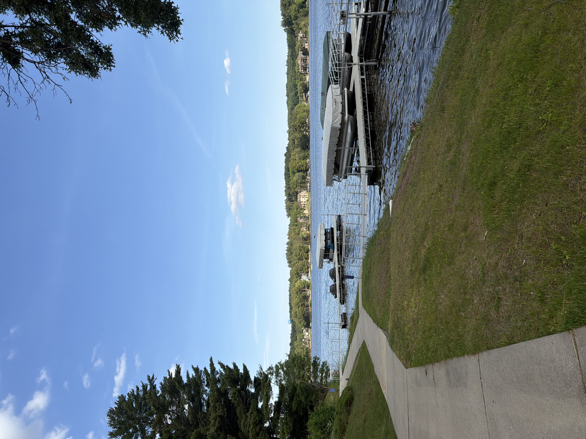 This image captures a serene daytime view of Lake Delton, United States of America. The scene is dominated by a clear blue sky with a few scattered white clouds, suggesting good weather.
In the foreground, a paved sidewalk curves gently along a lush, green grassy embankment that slopes down towards the lake on the right side of the frame. To the left, several dark green trees with dense foliage are visible, framing the view.
The midground features the calm, rippling water of the lake, stretching horizontally across the image. Several boat docks and lifts extend from the grassy shore into the water, with multiple boats, mostly covered, resting on these lifts.
The distant shoreline, visible across the lake, is covered in a dense line of green trees. Through the foliage, hints of residential structures or houses can be seen nestled among the trees. The overall impression is one of tranquility and natural beauty on a bright day. No discernible text is visible within the image.