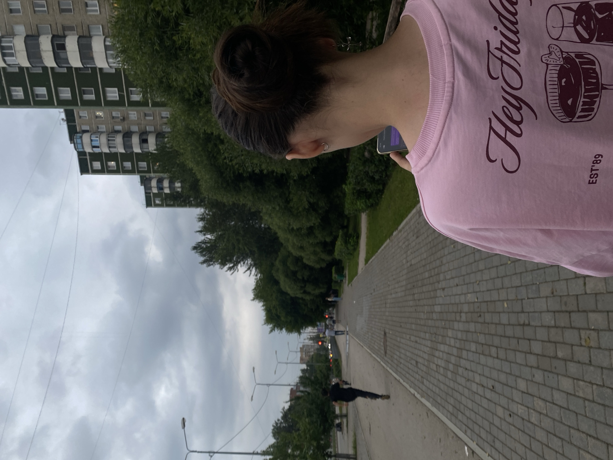 The image shows a woman from behind, wearing a pink sweatshirt with the text "Hey Friday" and "EST'89" on it, along with graphics of drinks. She is holding a smartphone. She is standing on a paved sidewalk. To her left, there is a dirt path and a road with cars and streetlights. Several people are walking or running on the path and sidewalk. In the background, there are trees and tall residential buildings. The sky is overcast. The scene is likely a street or park in a city. The woman's jewelry includes a hoop earring in her left ear and a cross-shaped earring in her right ear.