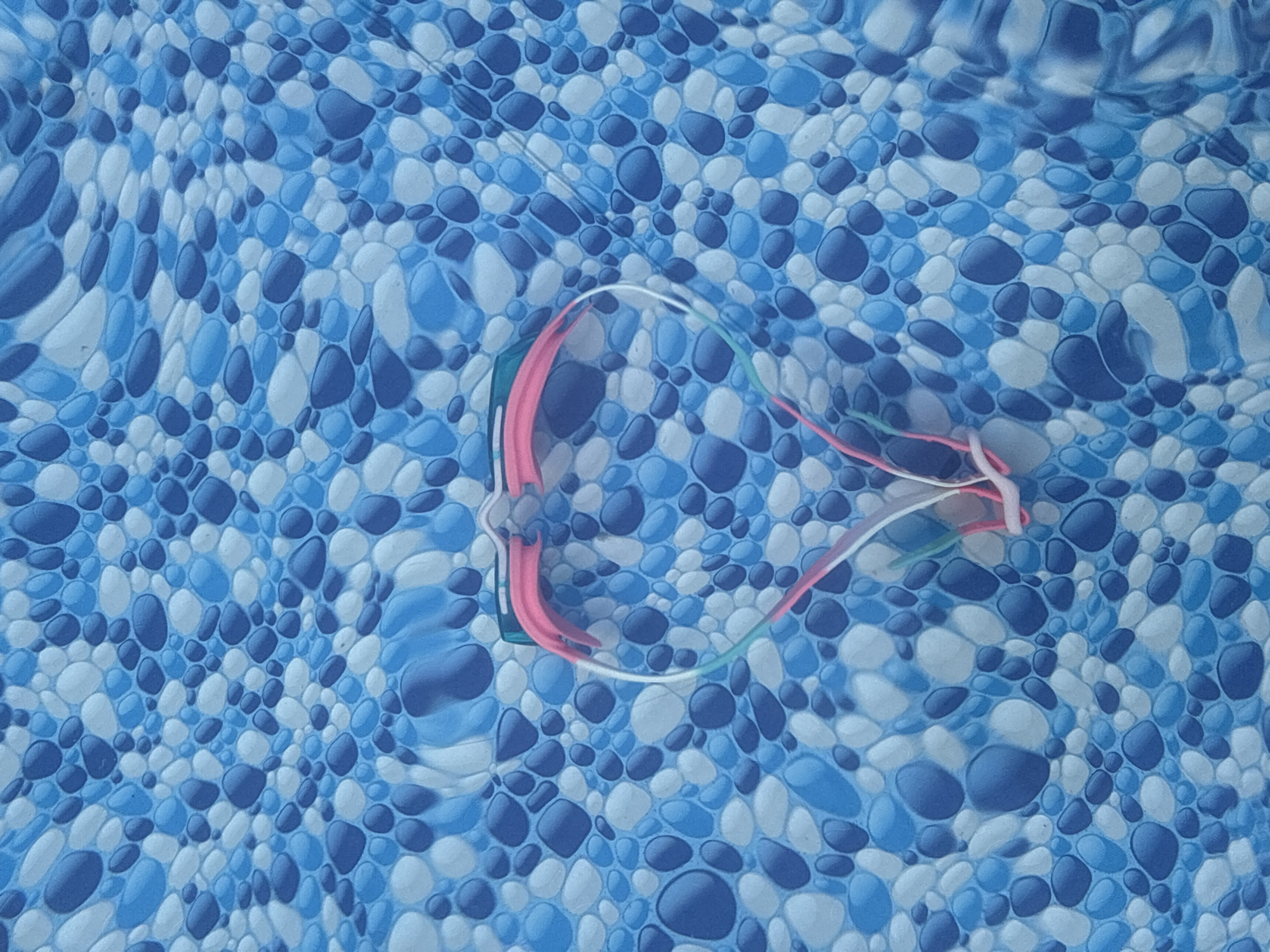 The image shows a close-up, overhead view of a swimming pool liner with a pebble-like pattern in shades of blue and white. A pink, white, and green swimming goggle strap is placed in the center of the frame, forming a heart shape. The goggle strap appears to be made of plastic and elastic. The water in the pool creates slight ripples and distortions on the liner. The image does not contain any information that would allow for the identification of the location in the city of Surami, Georgia.