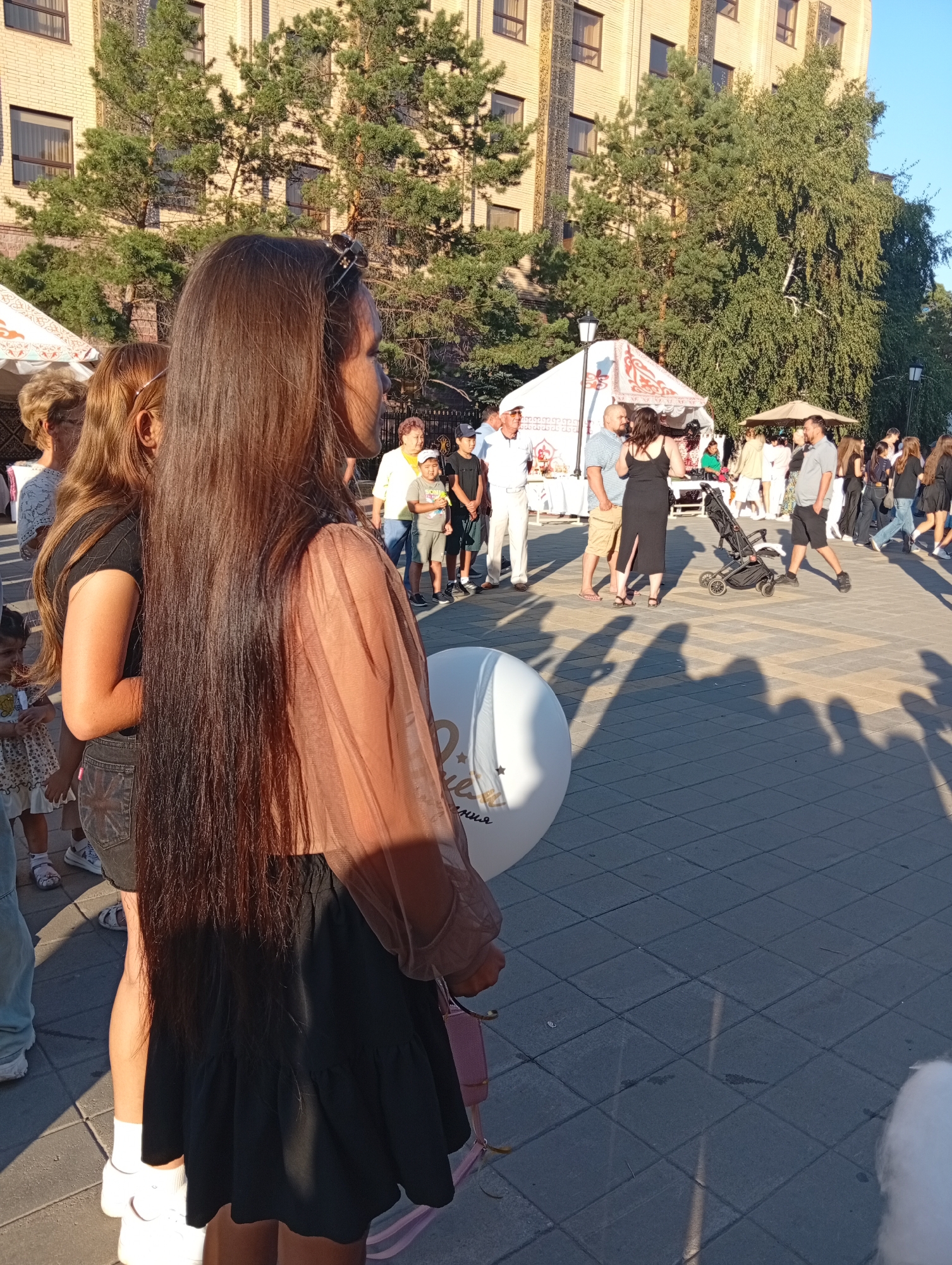 The image depicts a street scene, likely in Kostanay, Kazakhstan, on a sunny day. A young woman with long, dark brown hair is prominently featured in the foreground, facing right in profile and holding a large white balloon. She is wearing a sheer, light brown top and a black ruffled skirt. To her left, another woman with blonde hair is partially visible, also looking away from the camera.
In the background, a crowd of people are gathered, suggesting an outdoor event or market. There are several stalls with white tents and various items displayed. People of different ages are seen walking, talking, and interacting. A stroller is present near the center of the background. The architecture of a brick building with windows is visible behind the trees. The ground is paved with square tiles. The overall impression is of a lively public gathering.