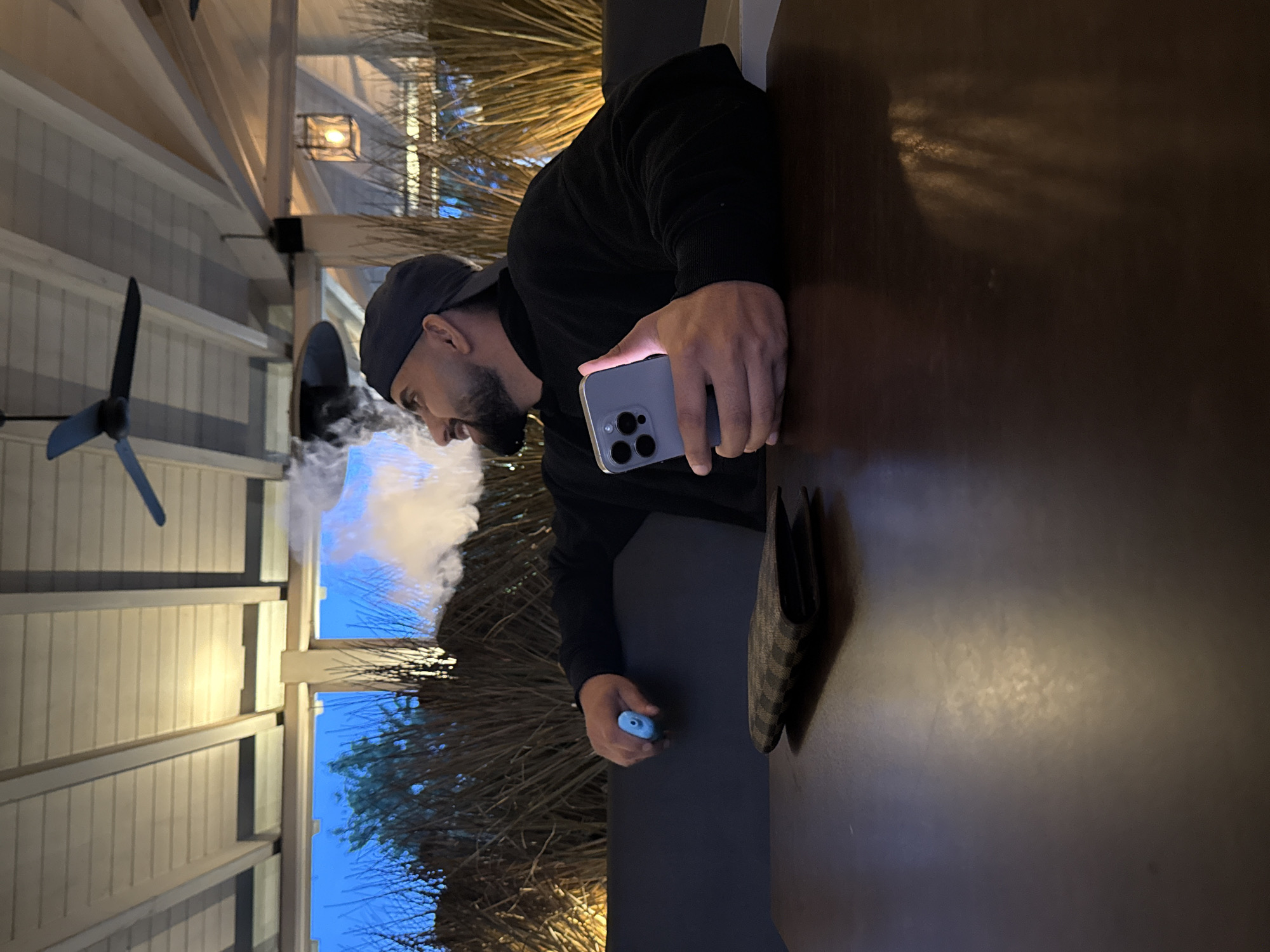 The image, rotated 90 degrees clockwise, captures a man vaping outdoors during what appears to be dusk or early evening in Gelsenkirchen, Germany. The man, seen in profile, is wearing a dark cap backwards and a dark long-sleeved top. He has a beard and is exhaling a large cloud of white vapor from a light blue vaping device held in his right hand. His left hand holds a modern smartphone, likely an iPhone, with its screen illuminated, resting on a dark tabletop. A dark, checkered wallet is also visible on the table next to his right arm.
The setting appears to be a covered outdoor patio or veranda, featuring white slatted walls and ceiling, supported by wooden beams. A dark ceiling fan with two blades is visible on the left, and a lantern-style light fixture hangs from one of the beams on the right. Beyond the covered area, the sky is a deep blue, suggesting the transition from day to night. Bare branches of trees or bushes are visible against the sky, indicating natural surroundings. The overall atmosphere is casual and relaxed. No visible text is present in the image.