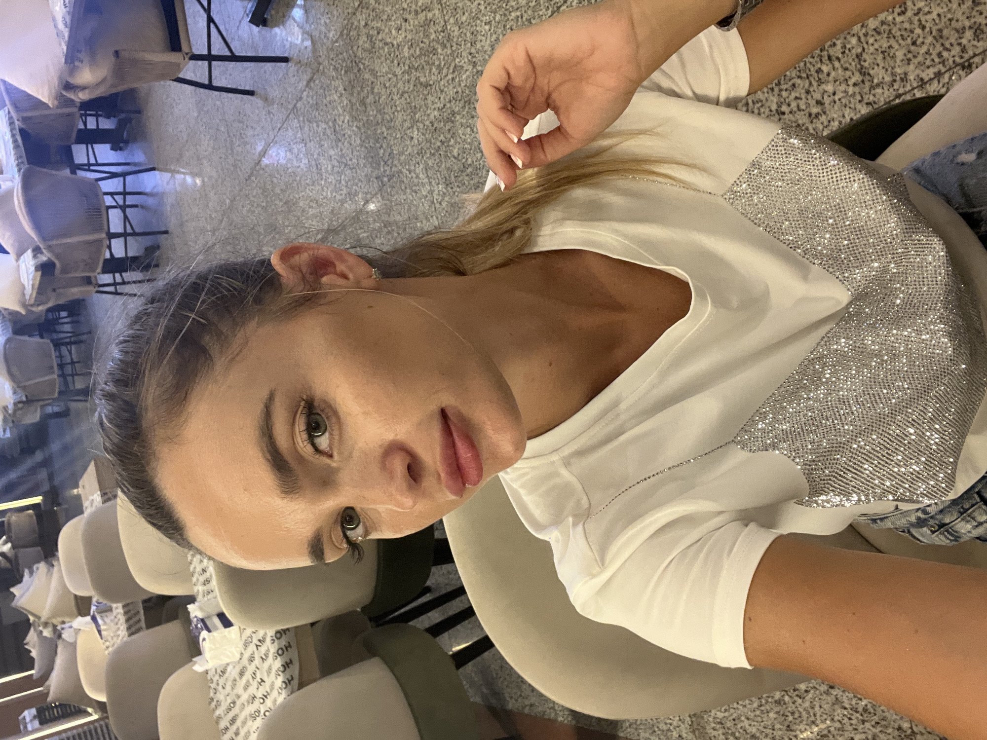 A woman with blonde hair pulled back in a ponytail takes a selfie in a well-lit, indoor setting that appears to be a cafe or restaurant. She is wearing a white t-shirt with a shimmering, silver, V-shaped embellishment across the chest, and denim shorts. Her makeup is visible, with emphasis on her eyes and lips. Her right hand is raised to her face, touching her chin. The background shows rows of empty chairs and tables, some with patterned tablecloths, suggesting a quiet time. The flooring is speckled. While the prompt mentions Sharm El Sheikh, Egypt, this is not visually verifiable from the image alone.