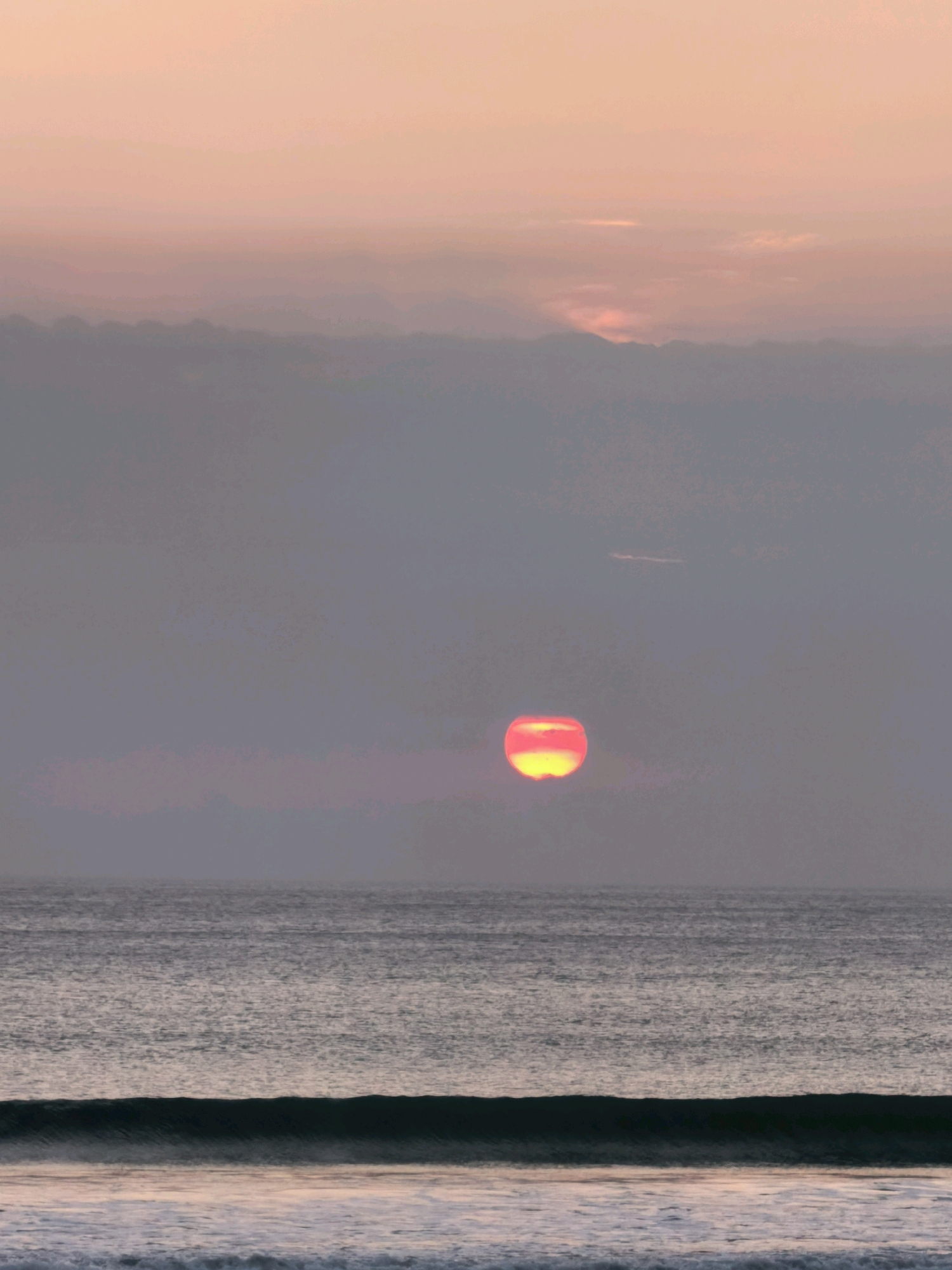 The image depicts a serene ocean sunset in Kuta, Indonesia. The sky is a gradient of soft pinks and oranges, transitioning to a muted grey towards the horizon. A large, vibrant orange sun is partially obscured by hazy clouds, casting a warm glow on the scene. The ocean in the foreground is calm, with gentle waves lapping at the shore. The water reflects the colors of the sunset, creating a shimmering surface. The overall impression is one of tranquility and natural beauty.