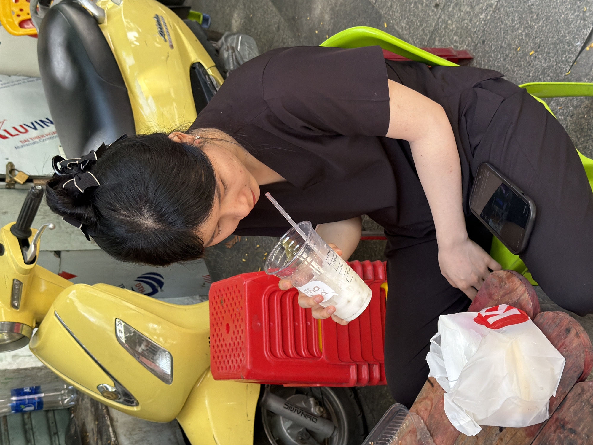 An Asian woman with her hair in a bun tied with a black ribbon is sitting on a bright green plastic chair next to a stack of red plastic stools. She is wearing a black V-neck top and black pants. She is holding a clear plastic cup with a straw in her right hand, appearing to drink from it. In her lap, to her left, is a white plastic bag with red lettering. A black smartphone rests on her right thigh. In the background, a yellow scooter is parked. The scene appears to be outdoors, possibly on a street or sidewalk in Nha Trang, Vietnam, given the presence of the scooter and the general ambiance.