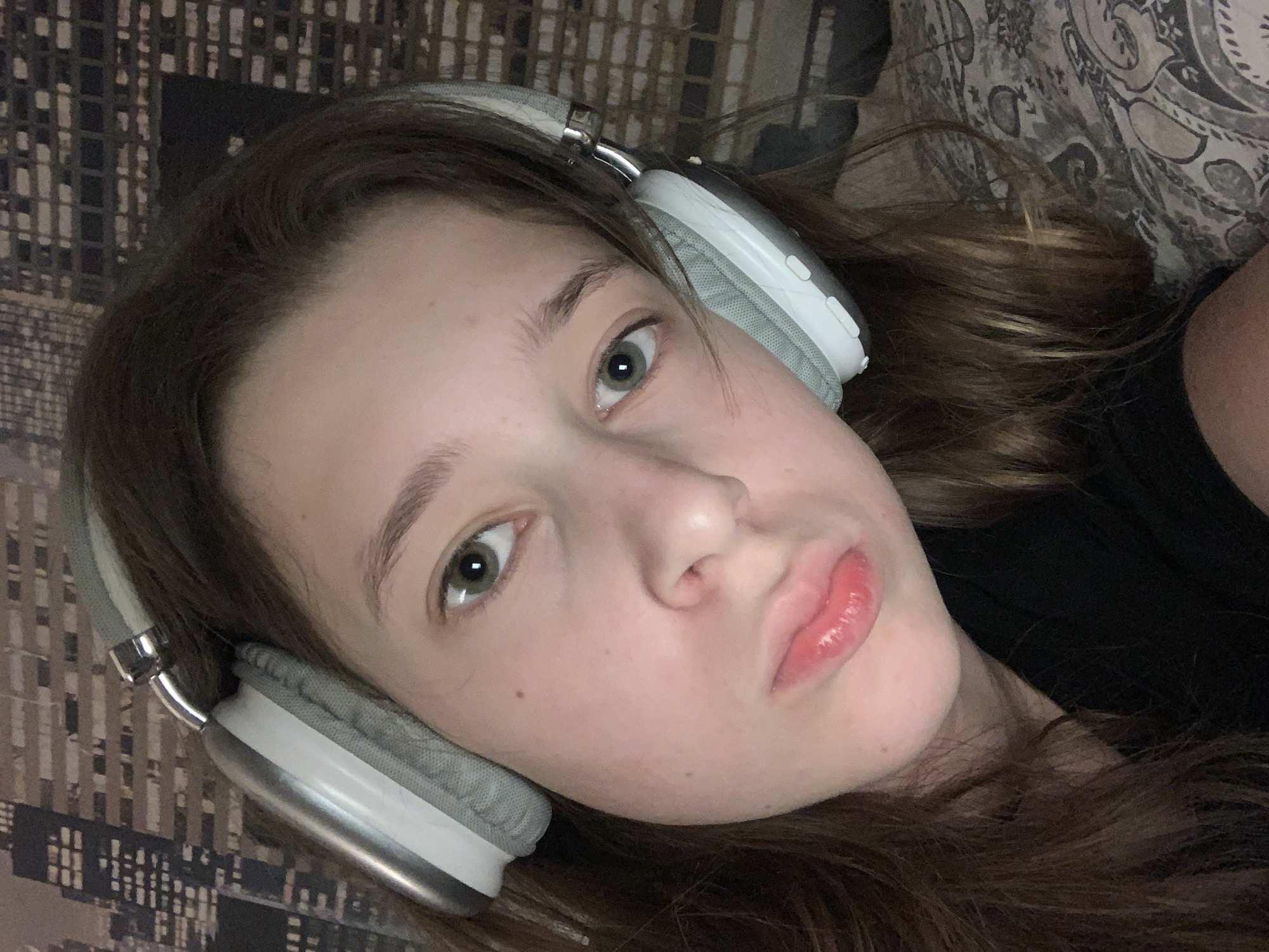 A young woman is lying down, wearing gray and white headphones. Her hair is brown with blonde highlights. She has fair skin, green eyes, and pink lips. She is wearing a black shirt. The background shows a patterned surface, possibly bedding or a blanket. The location cannot be determined from the image.