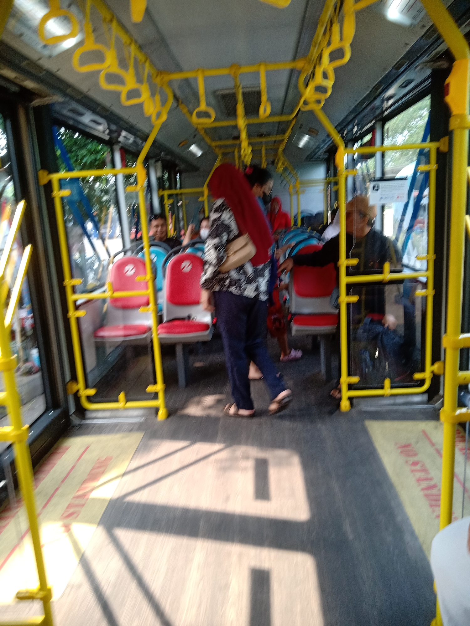 The image depicts the interior of a public bus in East Jakarta, Indonesia. The bus is yellow and grey, with red seats and yellow handrails. Several passengers are visible, some seated and some standing. The floor of the bus is grey with yellow and red markings, indicating a "no standing" zone. Sunlight streams through the windows, casting shadows on the floor.