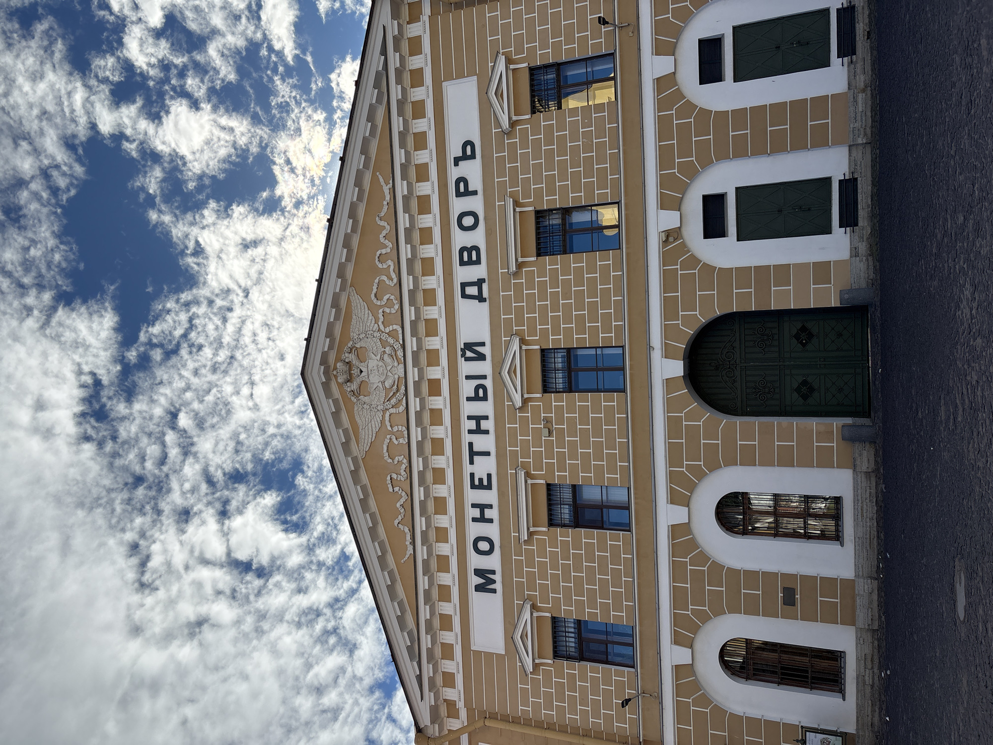 The image shows a building with the inscription "МОНЕТНЫЙ ДВОРЪ" in large, dark letters on a white horizontal panel. This inscription translates to "Mint Yard" or "Mints Court." The building is painted in a warm, yellowish-orange color with white trim, and it has a pediment at the top featuring a relief of a double-headed eagle with crowns, wings spread. Below the inscription are several arched windows with dark bars, and a large, ornate green double door in the center of the facade. The building's facade is constructed of bricks, with white lines outlining the brickwork. The ground in front of the building is paved with dark cobblestones. The sky above is blue with scattered white clouds.
This building is the Saint Petersburg Mint (Monetny Dvor), a historic mint located in Saint Petersburg, Russia. It is one of the oldest mints in Russia, founded in 1724.