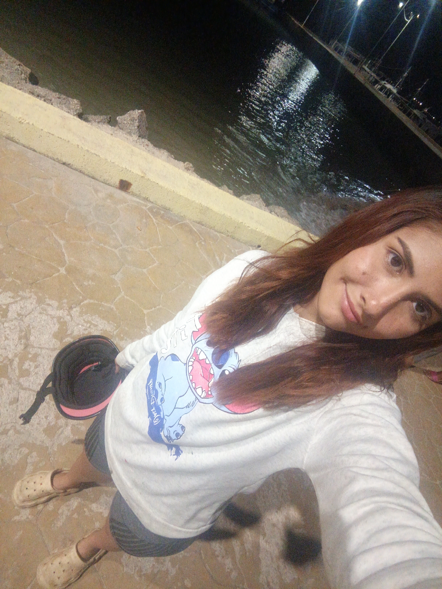 A young adult female, of Latin descent, with long reddish-brown hair, is captured in a high-angle selfie at night. She has a slight smile and is looking directly at the camera. She wears a white long-sleeved shirt featuring a blue cartoon character (Stitch) print, dark striped shorts, and light-colored slip-on clogs. A black object with a pink rim, possibly a helmet or bag, rests on the paved ground beside her left foot. The ground beneath her is a light-brown paved surface with irregular, interlocking tile patterns. To her right and behind her is a low, yellow or beige painted concrete wall, below which rough stone structures descend into a dark body of water. The water reflects multiple artificial light sources, creating shimmering patterns, and appears to be a bay or a large harbor. In the distance across the water, indistinct structures or poles with lights are visible, indicating a waterfront or pier environment. The scene is located in Juan Griego, Venezuela.