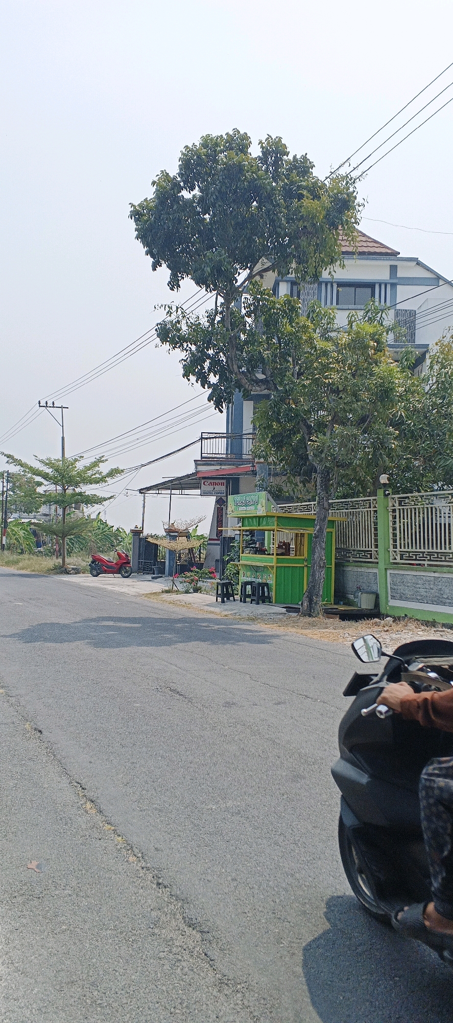 The image captures a street scene in Sonoadi, Indonesia, on a bright, overcast day. A gray asphalt road occupies the foreground, with a thin yellow line marking the lane division. A dark-colored motorcycle, partially visible from the rider's perspective, is in motion on the right side of the road. The rider's right hand is on the handlebar, and their left leg and foot, clad in patterned trousers and a sandal, are visible.
On the left side of the road, a red motorcycle is parked. Behind the red motorcycle, a small green food stall with yellow trim is set up, featuring several black stools and some decorative elements. A tall, modern building with multiple levels, painted white and blue, stands behind the food stall, partially obscured by trees. Power lines crisscross the sky above the scene. To the left of the building, a utility pole with electrical wires is visible. Trees with lush green foliage are scattered throughout the image, adding to the natural element of the urban landscape. A white metal fence with decorative patterns runs along the edge of the property where the building and food stall are located. The overall environment suggests a typical roadside setting in a populated area.