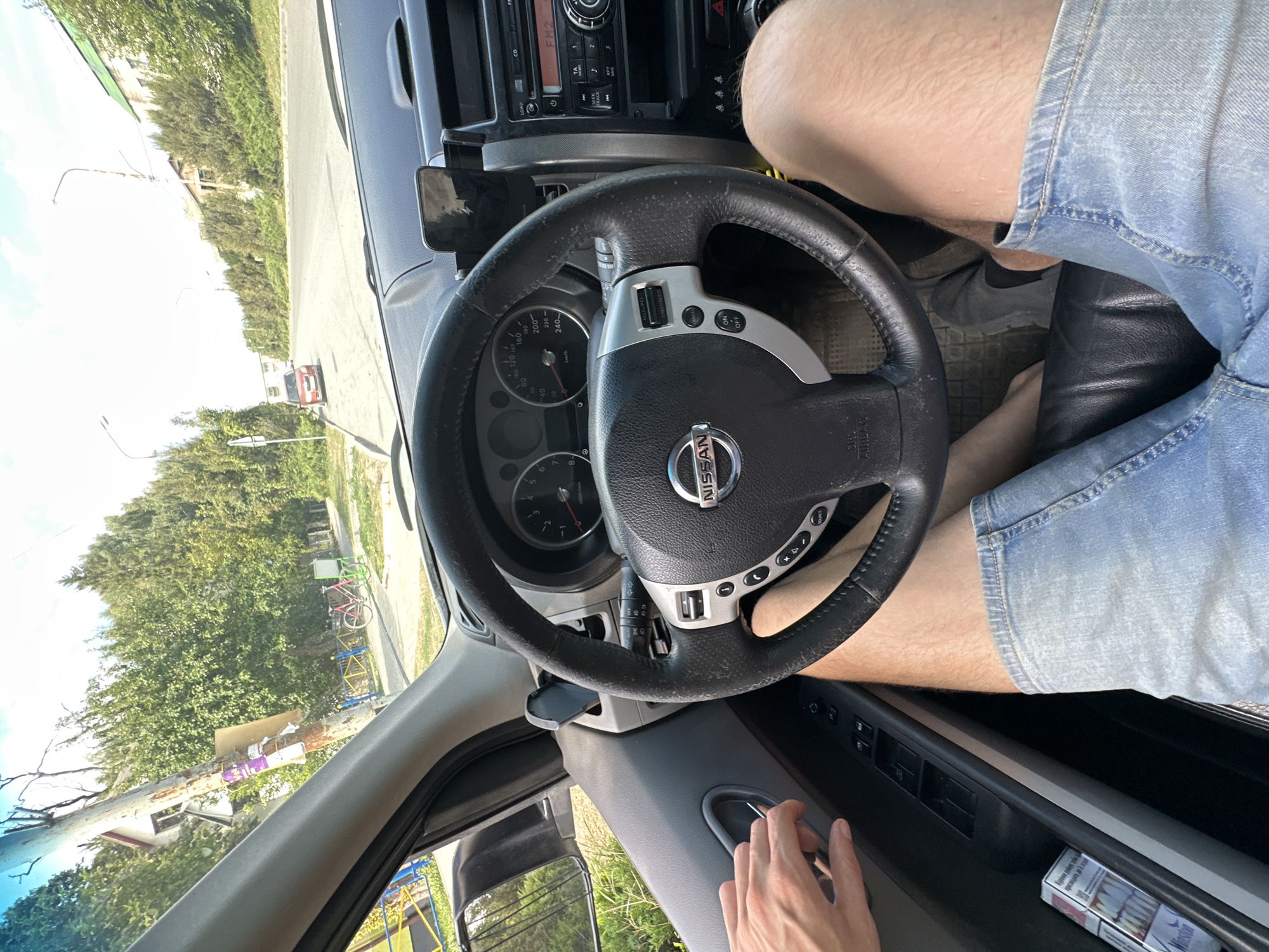 The image shows the interior of a Nissan X-Trail car, looking forward through the windshield. The steering wheel, dashboard, and a portion of the driver's legs are visible. The driver, wearing denim shorts, is seated in the driver's seat with their hand on the door handle. A pack of Winston cigarettes is visible in the driver's side door pocket. The exterior view shows a street with trees and buildings, suggesting an urban or suburban setting, likely Makiv, Ukraine, given the context. A red car is parked on the side of the road, and a bicycle is visible in the background. The sky is bright and clear, indicating daytime.