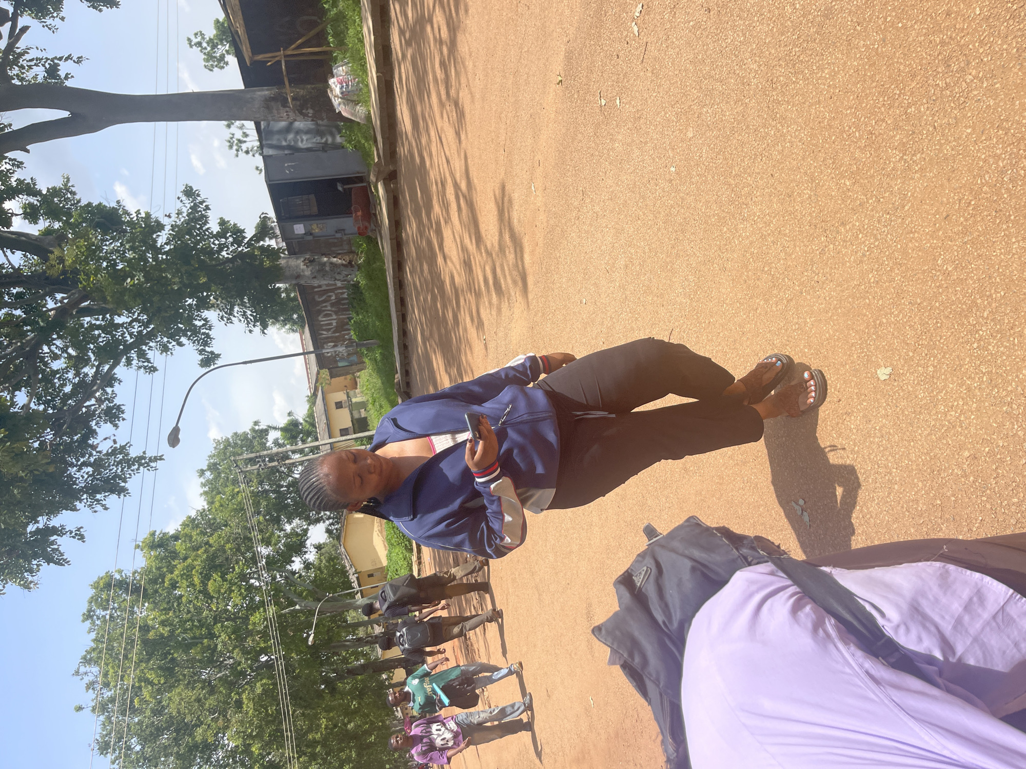 This media file captures an outdoor scene in Rafin Par, Nigeria. The central subject is a young woman standing on a dusty, reddish-brown path, holding a smartphone. She is wearing a blue athletic jacket over a white top and black leggings, with dark sandals on her feet. Her hair is styled in braids. Behind her, several other people are walking or standing, appearing to be students, some carrying backpacks or bags. They are dressed casually. The setting features a line of trees with lush green foliage on the left, and buildings in the background, including what appears to be a small shop or structure with graffiti on its wall. The ground is covered in a fine, granular material, common in arid or semi-arid regions. Overhead, power lines crisscross the sky, and a street light is visible. The presence of shadows suggests it is daytime, likely mid-morning or afternoon, with sunny weather. The overall atmosphere appears casual and everyday, possibly a school or university campus. There is visible text on the building in the background, appearing to be graffiti, including the word "KUDAS".