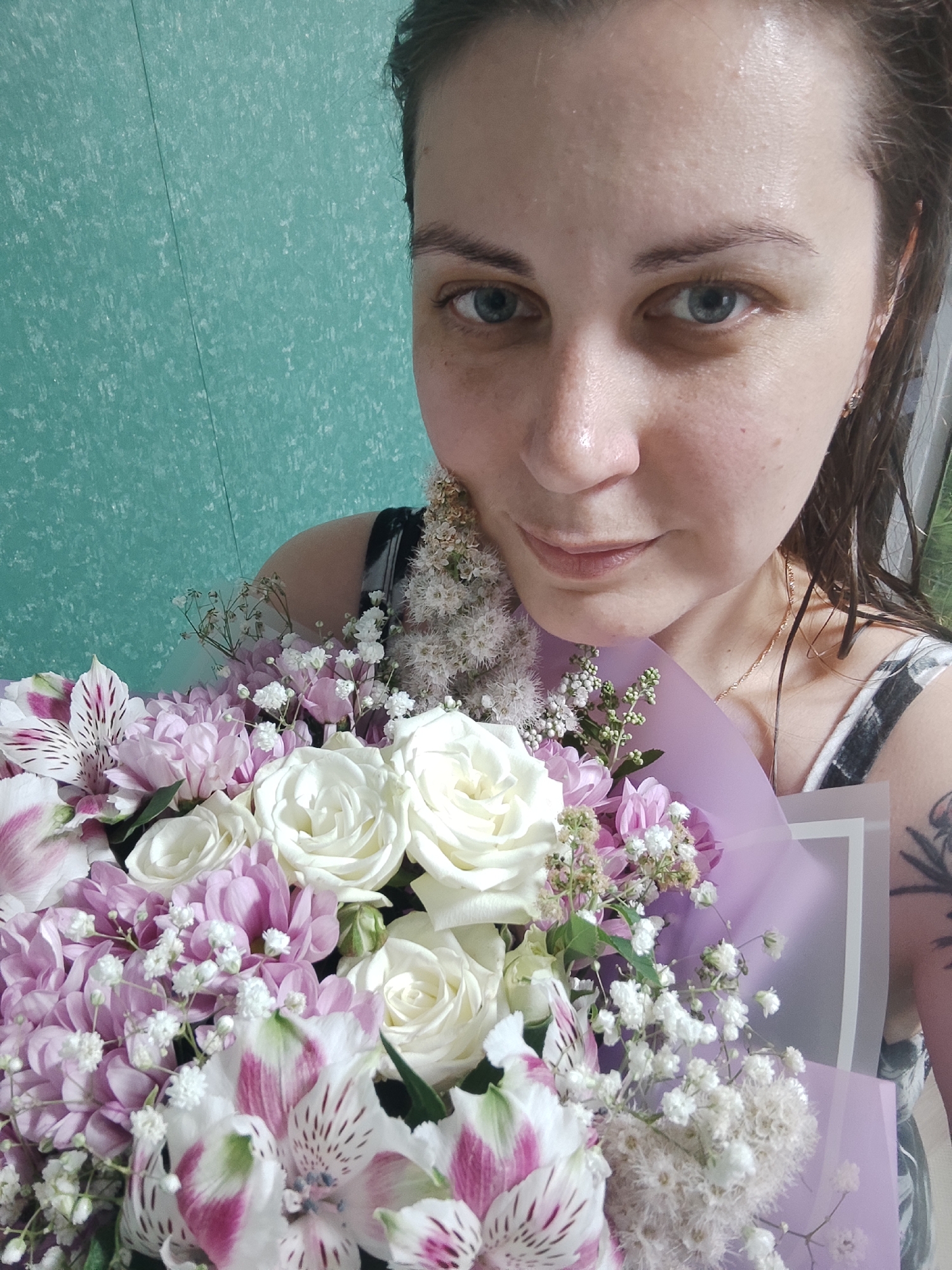 A close-up, high-angle, selfie-style shot shows a woman holding a bouquet of flowers in front of her face. The woman has light skin, blue eyes, and brown hair that is damp and pulled back. She is wearing a black and white patterned top. The bouquet contains white roses, pink chrysanthemums, white baby's breath, and pink and white alstroemeria. The background is a textured, teal-colored wall. The lighting suggests an indoor setting.