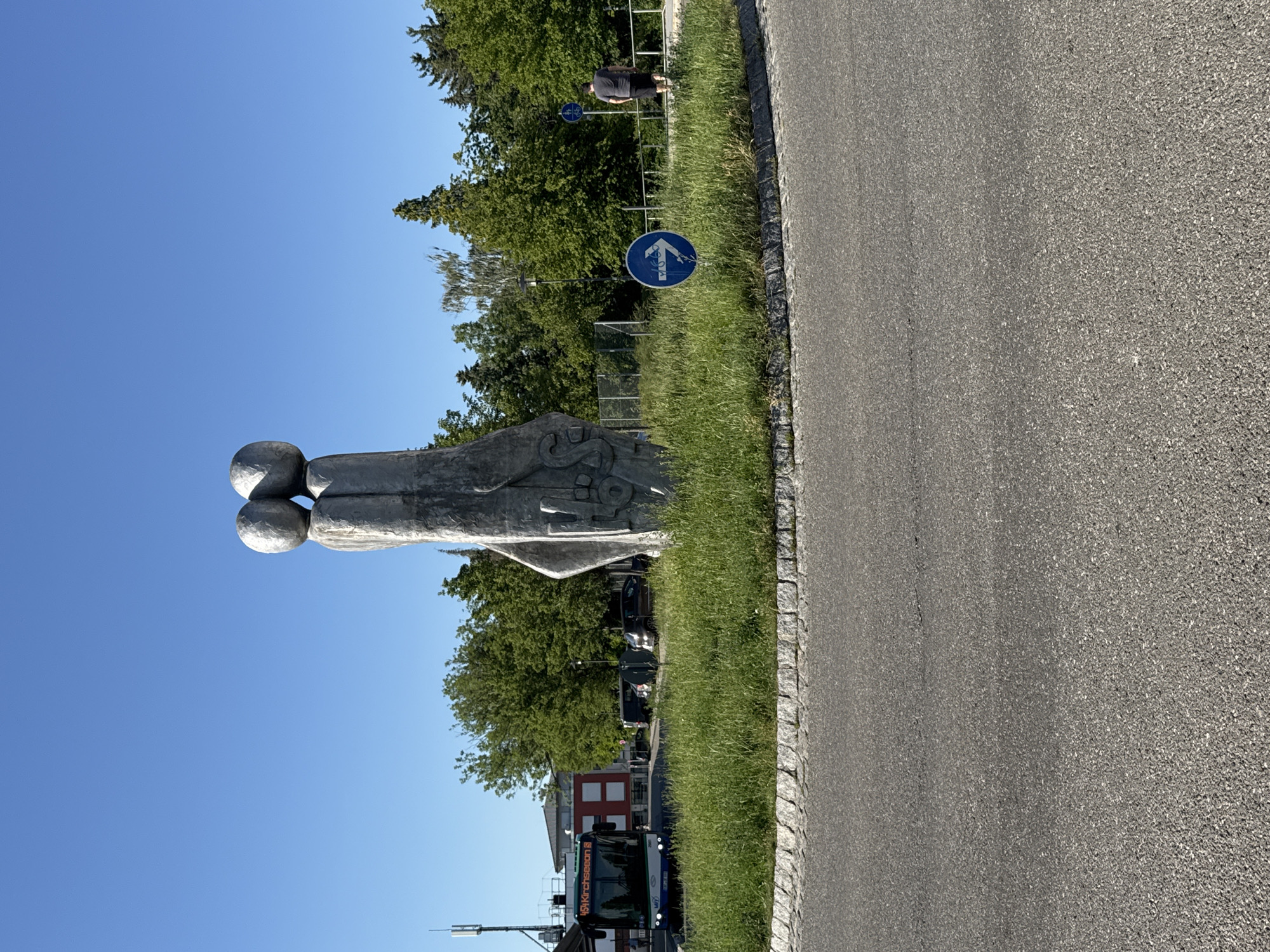 The image captures a scene in Höhenkirchen-Siegertsbrunn, Germany, on a bright, clear day, likely mid-afternoon due to the strong sunlight casting shadows. The foreground is dominated by an abstract sculpture made of metal, featuring rounded shapes that resemble spheres and organic forms. To the right, a paved road with a stone border runs alongside a strip of lush green grass.
In the background, a line of trees forms a dense green backdrop against a vibrant blue sky. Further beyond the trees, hints of urban life appear, including a red-brick building and what seems to be a bus. A blue circular traffic sign with a bicycle symbol and an arrow is visible on a post, suggesting a dedicated cycling path or route nearby. Another blue sign with a different symbol is also present.
A person is standing in the middle ground, observing the surroundings, dressed in dark clothing. The overall mood is peaceful and everyday, typical of a suburban or semi-urban environment. The sunlight highlights the textures of the sculpture and the road, and the clear sky indicates fair weather. The presence of a bus with visible text on its front suggests it is a public transport vehicle operating in the area. The text on the bus reads "194 Kirchseeon S".