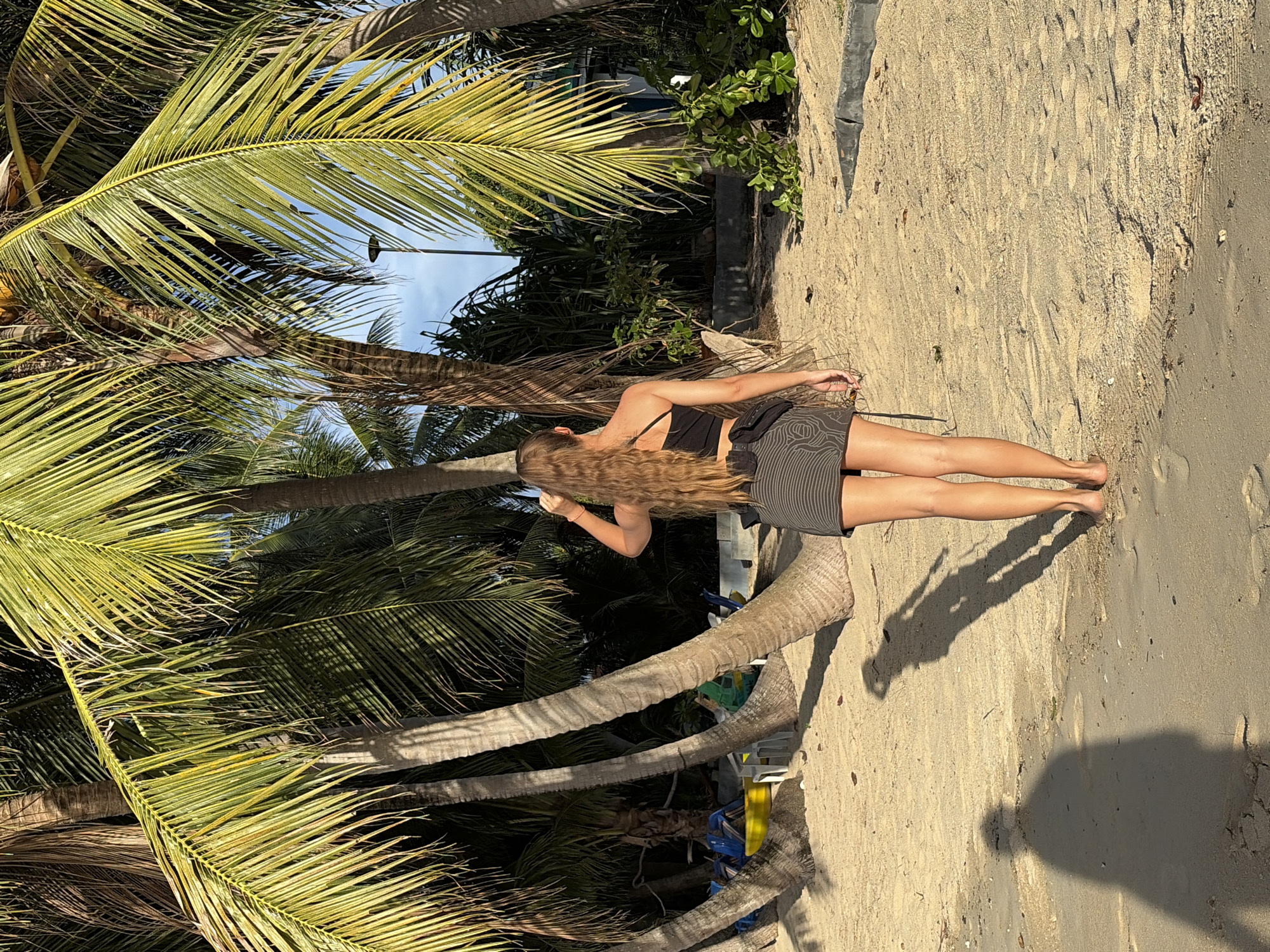 A woman with long, wavy brown hair stands on a sandy beach, facing away from the camera. She is wearing a black crop top and a short, gray and black striped skirt. Her left hand is raised, adjusting her hair. She is surrounded by palm trees, with their fronds casting shadows on the sand. In the background, there are some chairs and a building. The scene appears to be in Baan Thong Po, Thailand.