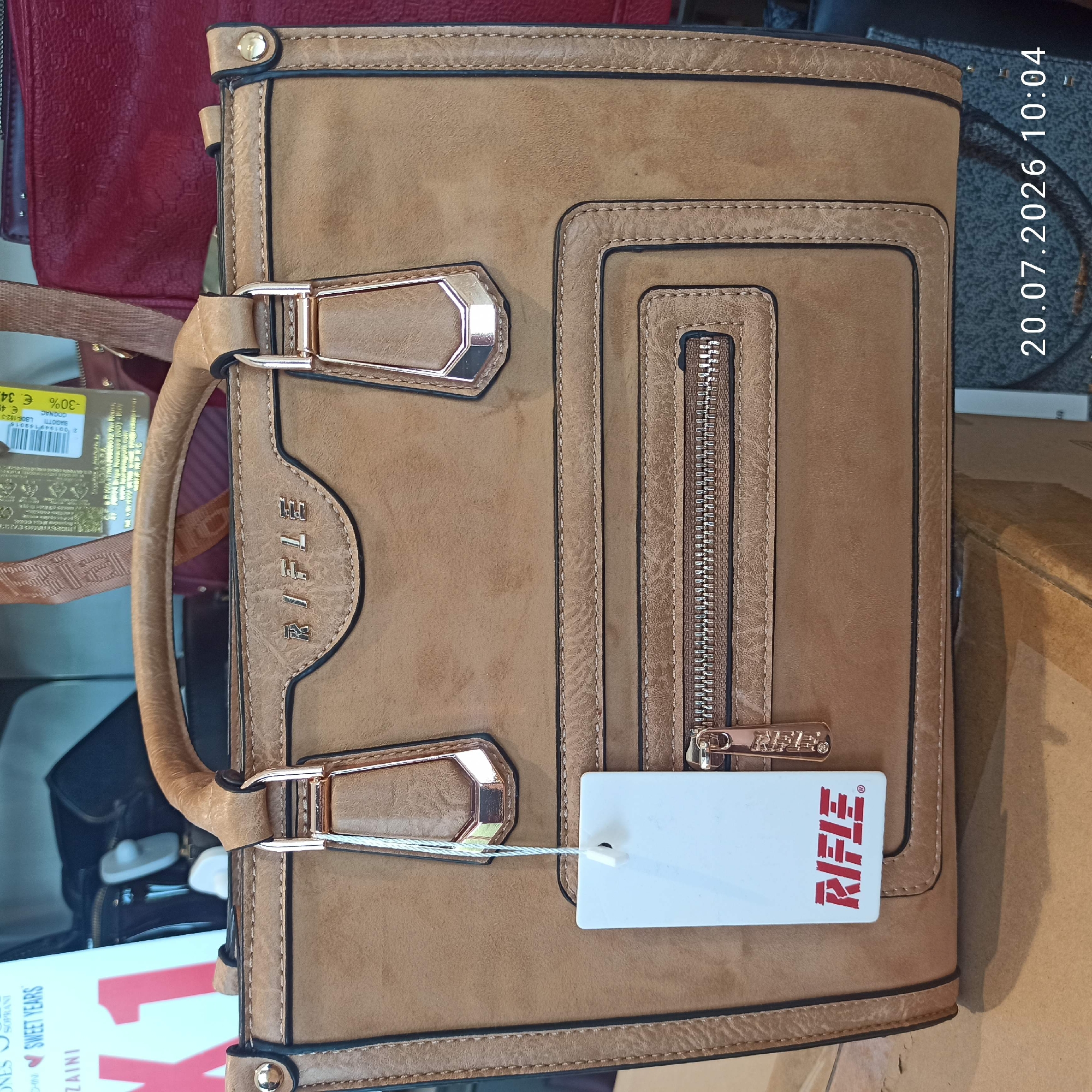 A light brown, structured handbag with gold-toned hardware is the main subject of the image. The handbag has a central zip pocket on the front, and two top handles attached by gold-toned clasps. A white tag with a red "RIFLE" logo hangs from the left handle. The bag is placed on a surface, possibly a shelf or a table, with other bags visible in the background. To the right of the handbag, a dark red textured bag is partially visible. The overall scene suggests a retail environment, likely a shop or boutique. The timestamp "20.07.2026 10:04" is overlaid on the bottom right corner of the image. The image was taken in Bari, Italy.
