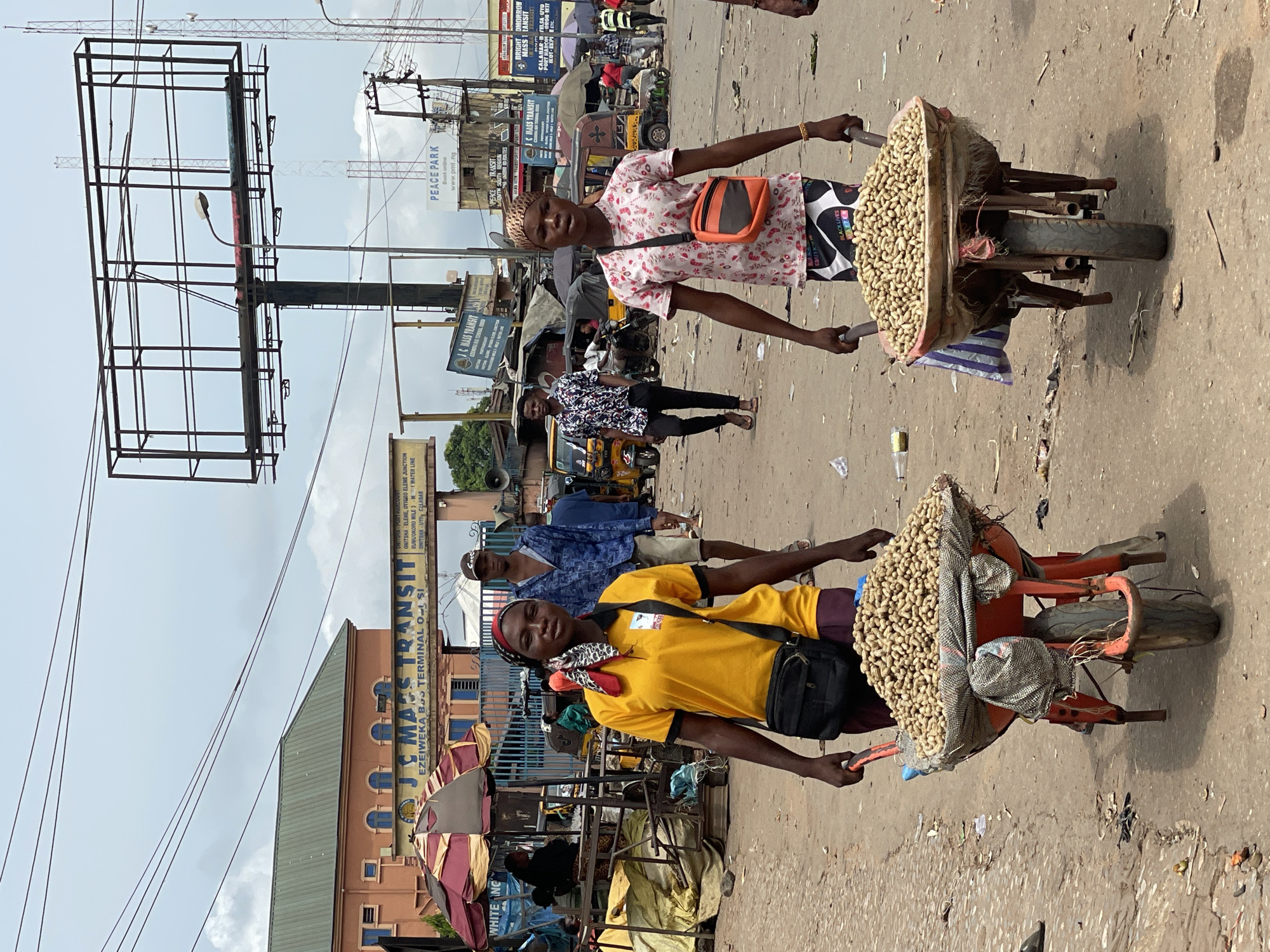 The image depicts a street scene in Nkpor, Nigeria. Two women are positioned in the foreground, each pushing a wheelbarrow laden with peanuts. The woman in the yellow shirt is closer to the viewer, followed by the woman in the pink patterned shirt. They are walking on a paved road with visible litter and debris. In the background, other pedestrians and vehicles are present. Signage includes "JC Mass Transit," "Peace Park," and "Bright Omorrow Mass Transit." An empty billboard frame is on the left. Power lines traverse overhead. The day appears overcast.