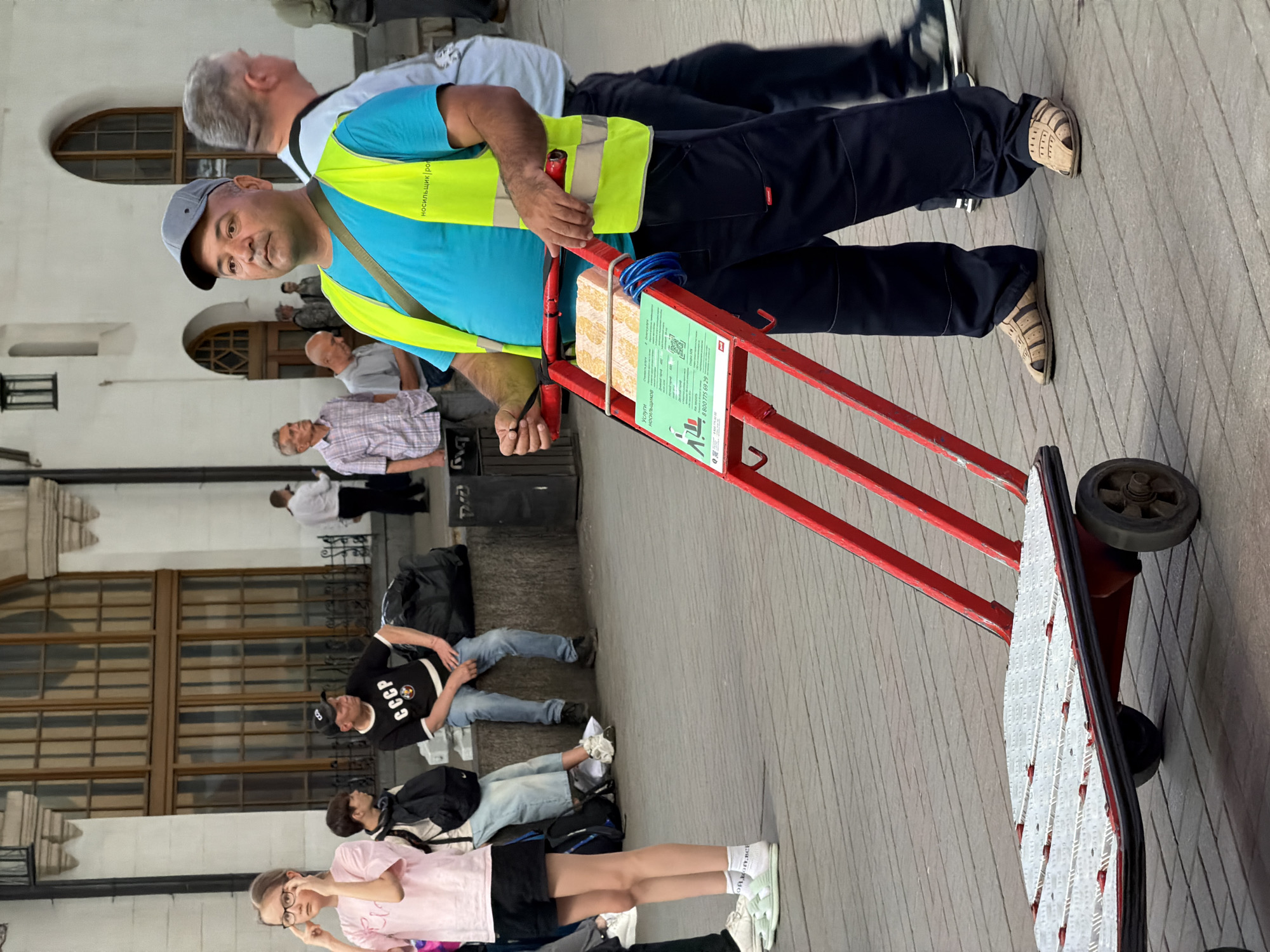 The image depicts a man pulling a red hand truck on a tiled sidewalk in what appears to be a public square in Moscow, Russia. The man wears dark pants, a light blue shirt, a yellow safety vest, and a gray baseball cap. He is looking at the camera with a serious expression. The hand truck has a sign written in Cyrillic script (Russian). The sign on the hand truck includes the text: Услуги носильщиков (Services of Porters) and a phone number.
Several other people are present in the background. Some are seated on the ground, while others are standing or walking by.
Buildings with arched windows are visible in the background. The overall scene suggests a busy public space, likely a train station or a similar transportation hub.