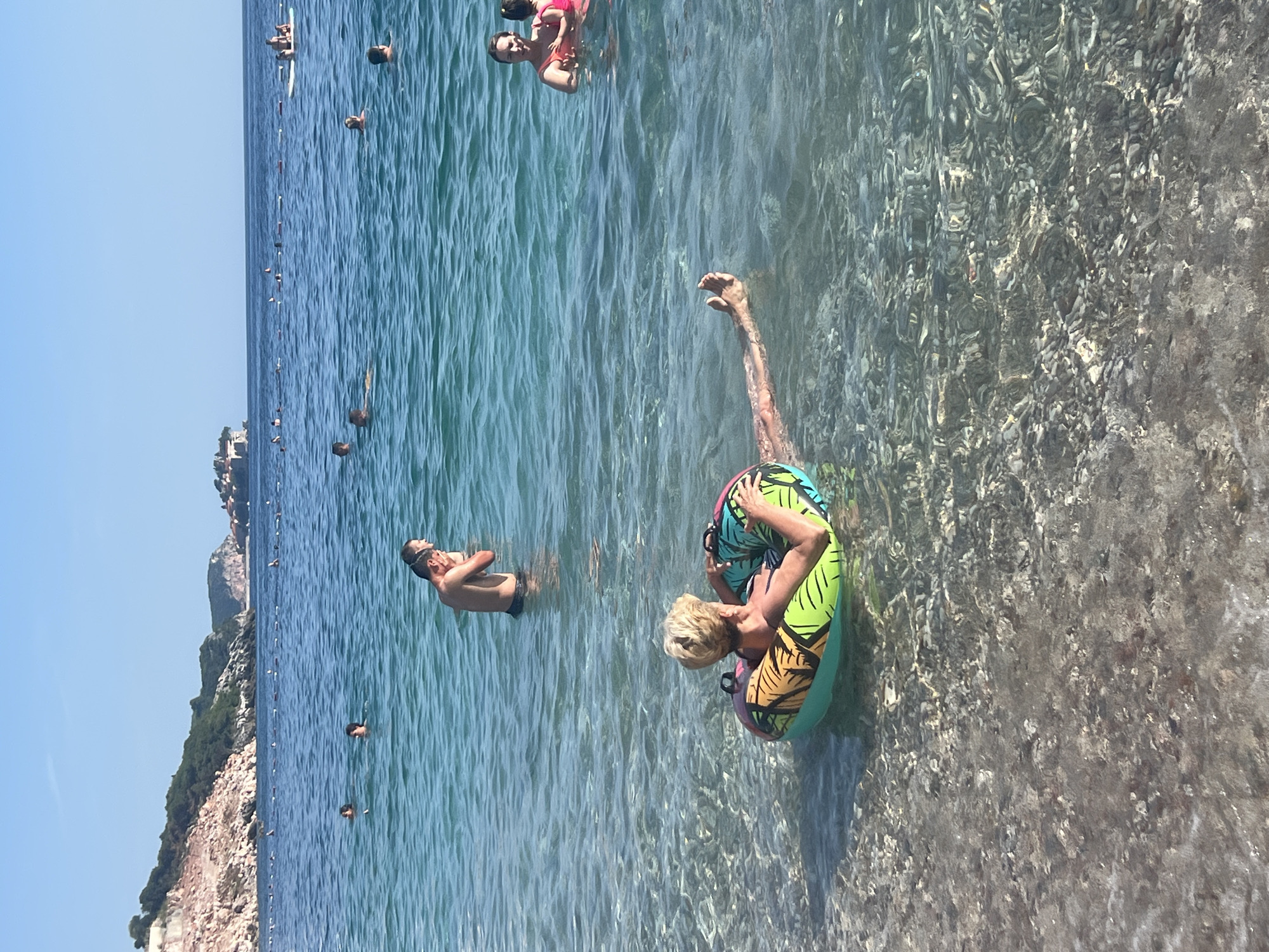 In Kamenovo, Montenegro, on a clear, sunny day, several people are enjoying the turquoise waters of the Adriatic Sea. In the foreground, a person with blonde hair floats on their back in a colorful inflatable ring, their legs extended straight out in front of them, submerged in the shallow, clear water. The seabed is visible beneath the surface, covered in small pebbles. Further out in the water, a man stands partially submerged, looking towards the shore. In the middle ground, more swimmers are scattered across the sea, and a group of people are on a paddleboard. In the background, the coastline is lined with rocky cliffs and green vegetation, with buildings visible in the distance. The sea is calm, with gentle ripples on the surface, and the horizon is clear under a bright blue sky.