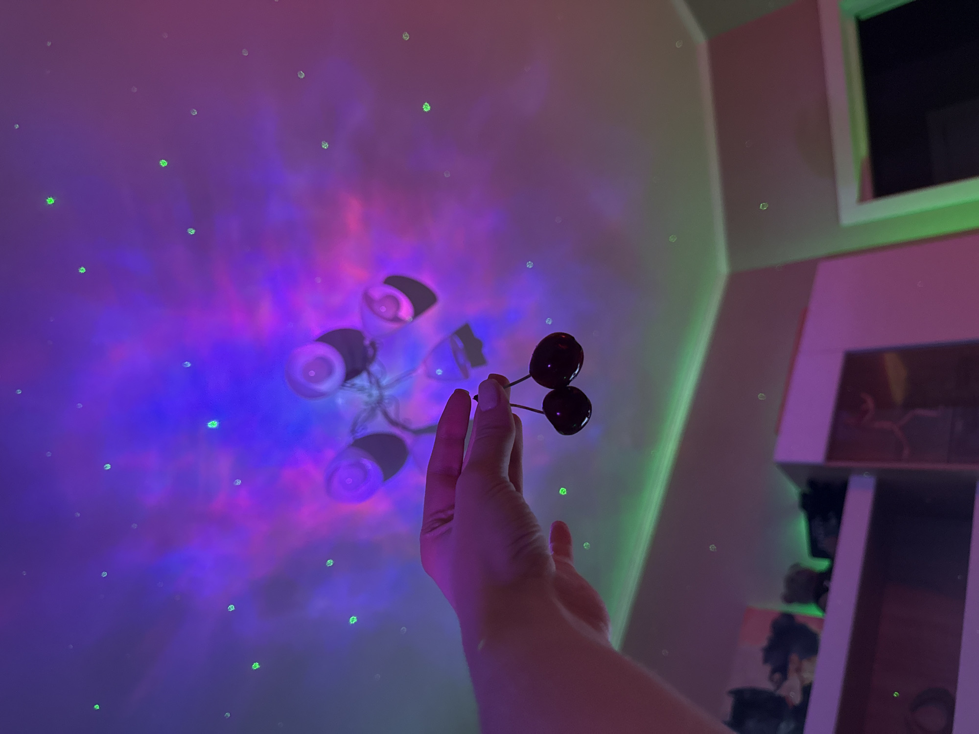 A person's hand is holding two cherries against a ceiling illuminated by a galaxy projector. The projector casts swirling patterns of purple, blue, and pink light, along with scattered green dots resembling stars. Green LED strip lights are visible along the wall and ceiling edges, and shelves with various objects, possibly toys, are in the background. The overall scene is dimly lit and atmospheric. The location is unknown, Unknown.