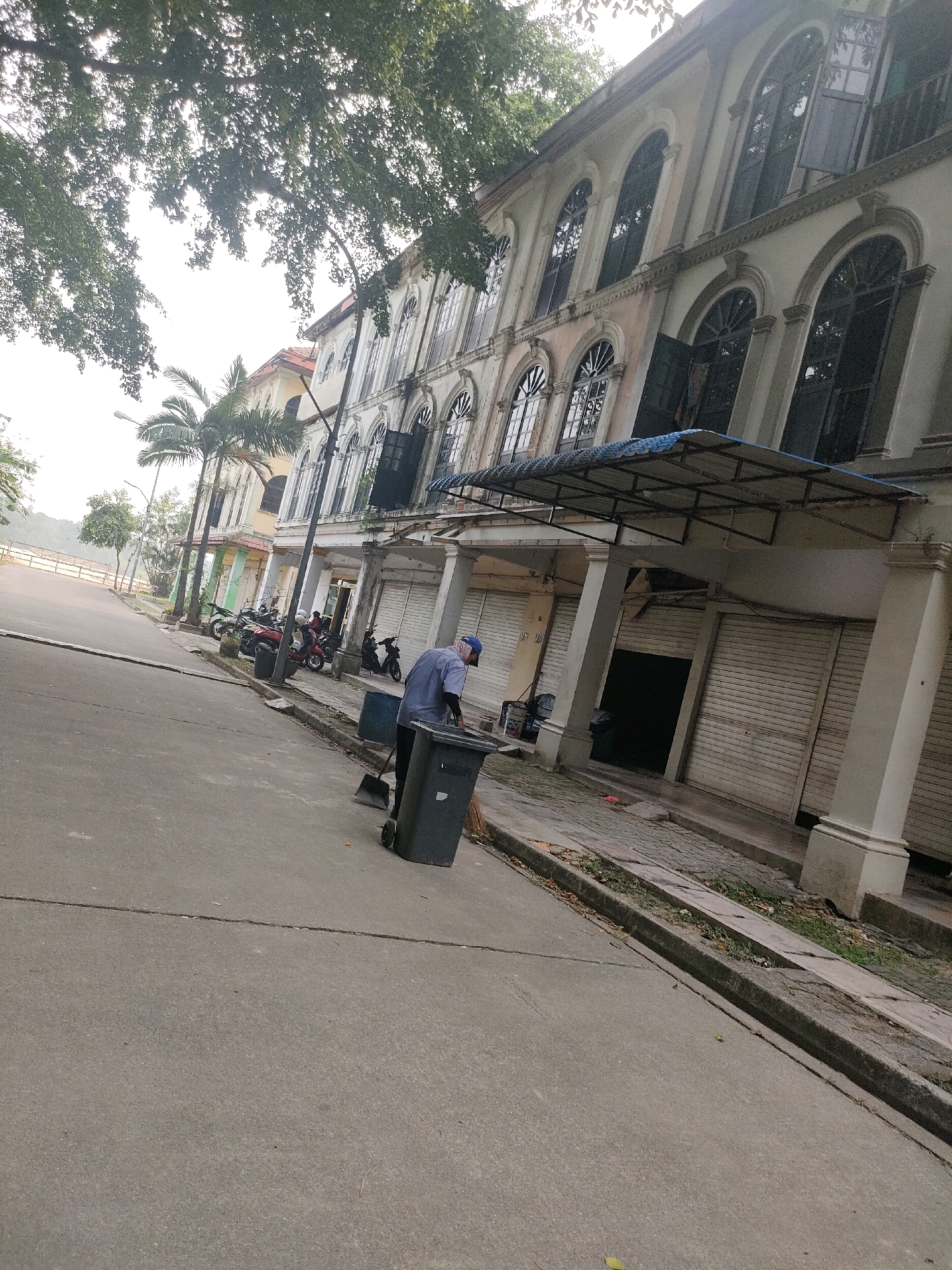 The image depicts a street scene in Batam, Indonesia, with a focus on a building with arched windows and several parked motorcycles. In the foreground, a person wearing a blue cap and a grey shirt is sweeping debris into a large grey garbage bin with a broom. The street is paved, and there is a concrete gutter alongside the sidewalk. The building is two stories high with a light-colored facade and multiple arched windows on the upper floor. The ground floor appears to be commercial with roller shutters. Palm trees are visible behind the building, and lush green foliage from trees overhangs the scene from the left. The sky is bright, suggesting daylight. The overall impression is of a quiet street, possibly in a commercial area of Batam.