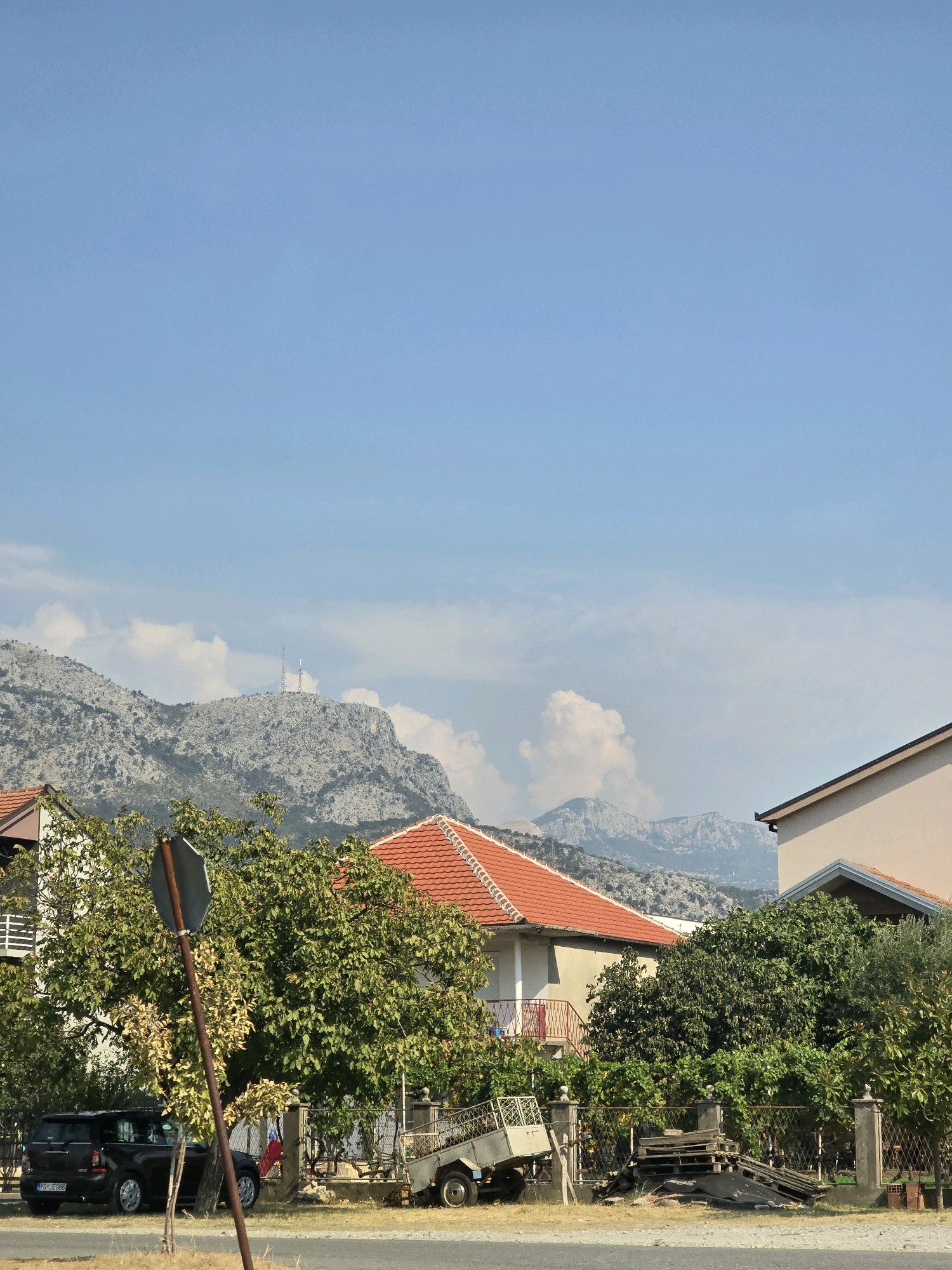 The image shows a bright, sunny day with a clear blue sky over a hilly landscape. In the foreground, there are several houses with red-tiled roofs, surrounded by lush green trees. A dark-colored car is parked on the left side of the image, and a small trailer is visible on the right. In the distance, a mountain range is visible, with two tall radio towers on its peak. The scene appears to be a residential area in a rural or semi-rural setting. The city of Podgorica, Montenegro is not explicitly depicted or suggested in the image.