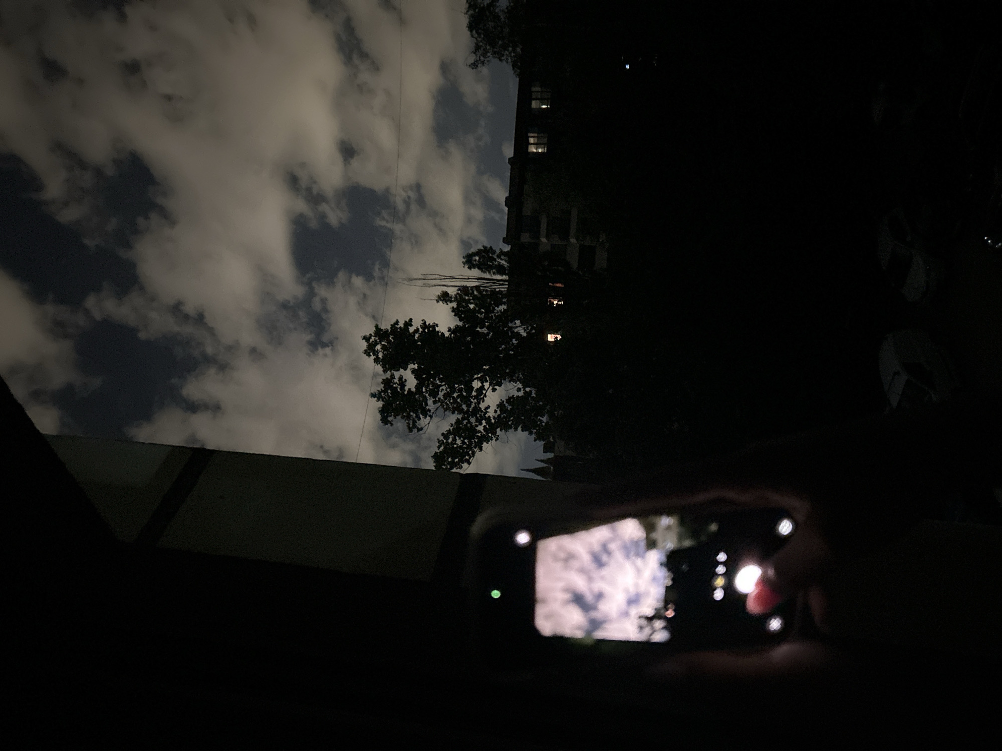 A nighttime shot, likely taken in Sofia, Bulgaria, showcases a person holding a smartphone to photograph the sky. The sky is filled with thick, luminous clouds against a dark background. The phone's screen displays a similar image of the sky, mirroring what is being photographed. In the background, silhouetted trees and the outline of a building with a few illuminated windows are visible. To the right, indistinct shapes could be vehicles or objects in the darkness. The foreground includes part of a roofline or building edge.