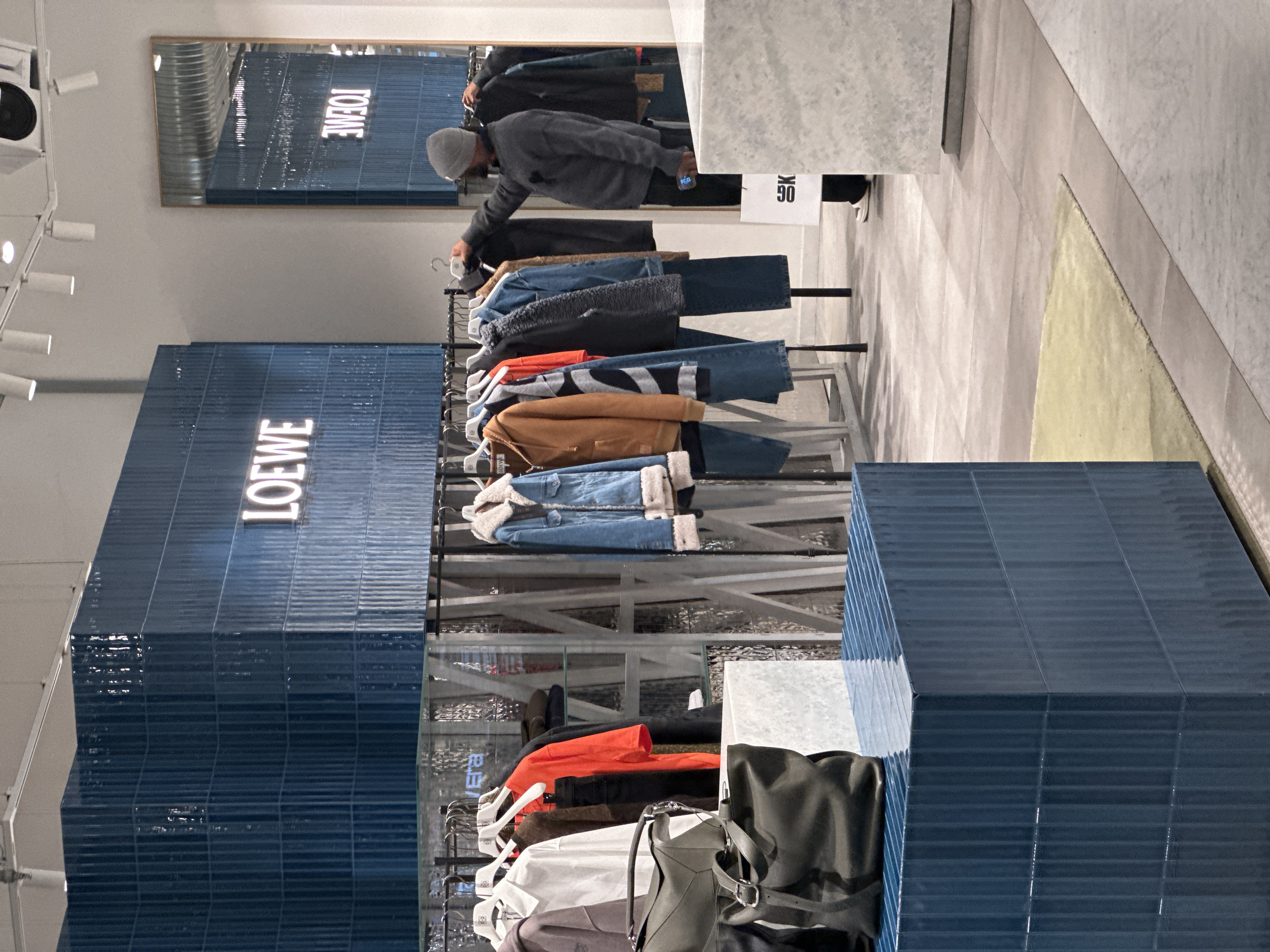The image depicts the interior of a Loewe luxury fashion store in London, United Kingdom. The central feature is a large, rectangular display structure constructed from dark blue, vertically ribbed ceramic tiles, bearing the illuminated "LOEWE" brand name in white block letters. This structure is positioned behind a black metal clothing rack.
On the right side of the frame, a lone individual, seen from behind, is examining clothing on the rack. This person wears a grey knit beanie and a dark grey long-sleeved garment, with a smartphone held in their right hand. A white shopping bag with partial black lettering is visible near their feet, resting against a light-colored plinth. The clothing rack displays a variety of garments, including a light blue denim jacket with a cream sherpa collar, a brown hooded sweatshirt, a white collared shirt, and an orange long-sleeved top, all suspended on white hangers, some bearing the Loewe logo.
In the foreground, a lower display stand, also clad in the dark blue ribbed tiles, holds two dark grey or black leather handbags. The store environment features light grey walls, a white ceiling with multiple track lights and an exposed white air conditioning unit with a black circular vent. A large, rectangular mirror with a wooden frame on the right wall reflects parts of the store, including the reversed "LOEWE" sign. The floor consists of light grey/beige rectangular tiles, with a segment of pale yellow carpet visible. Other display elements include transparent glass shelving.