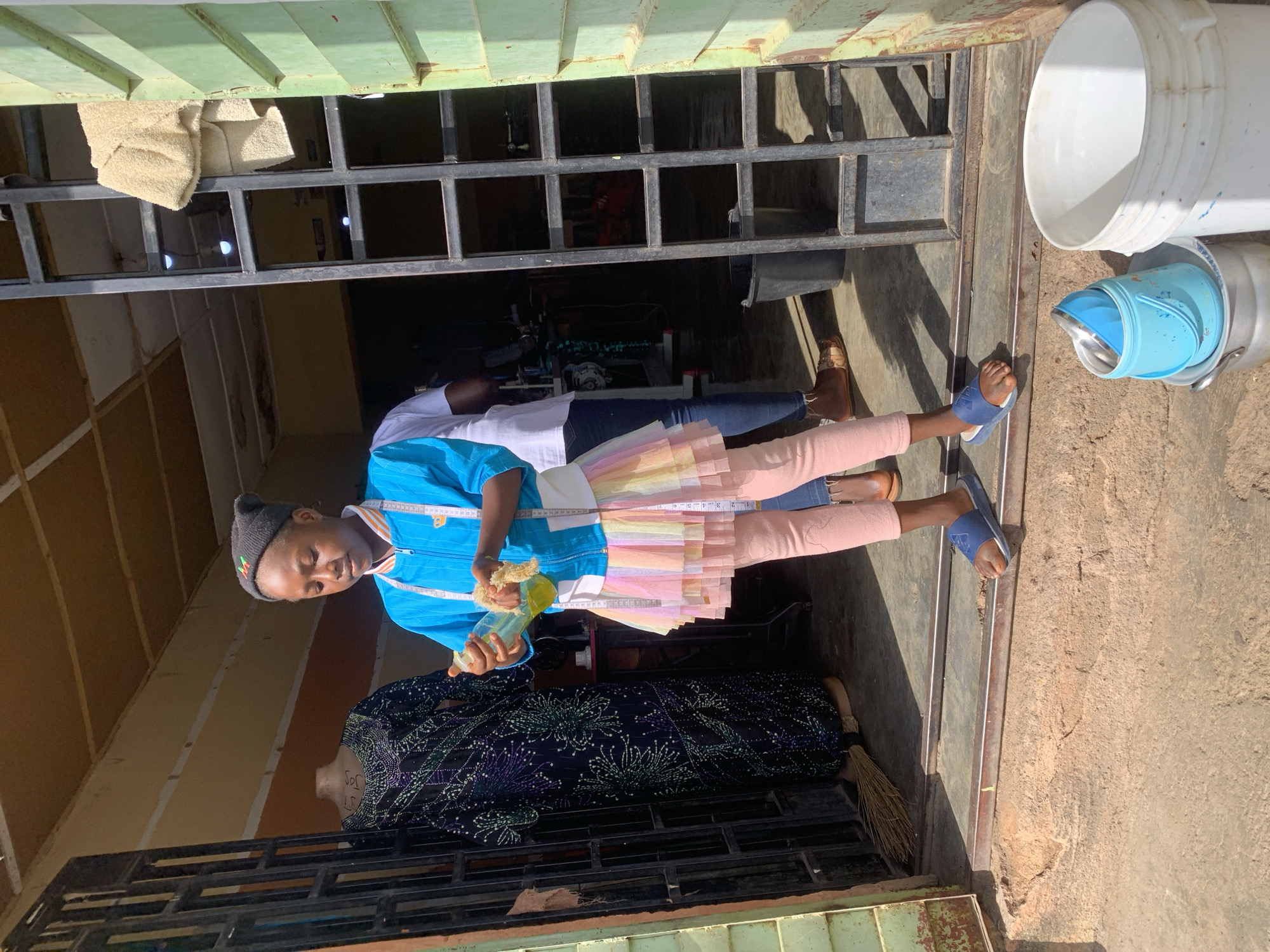 A person wearing a blue jacket, pink leggings, and a colorful skirt stands in the doorway of a building. A measuring tape is draped around their neck. They are holding a bottle in one hand and a sponge in the other. Behind them, there is a sewing machine and a mannequin draped with patterned fabric. Outside the doorway are stacked white and blue buckets. The location is likely Jos, Nigeria, based on the subject's appearance and surroundings. The environment appears to be a workshop or storefront.