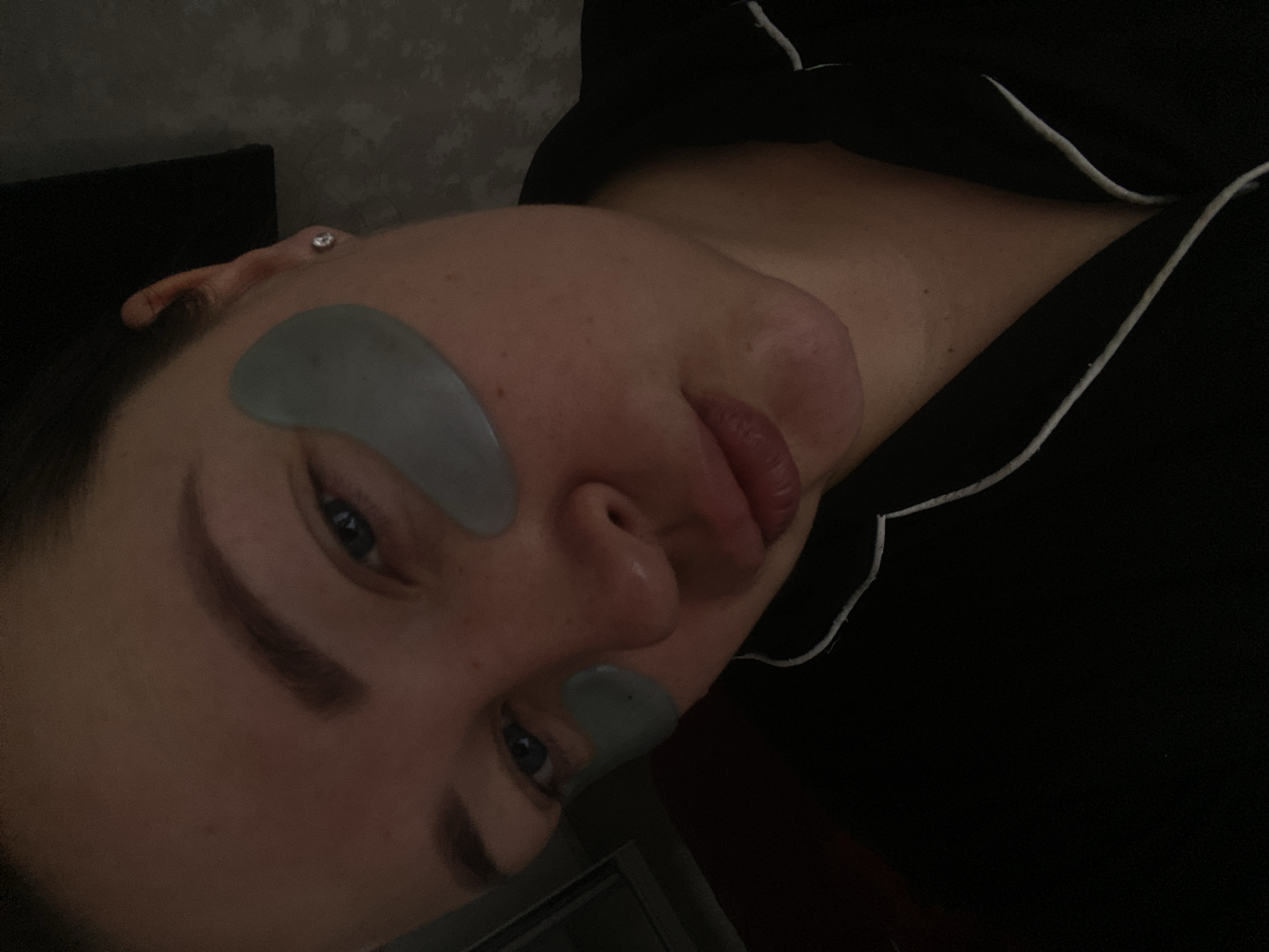 A female individual is depicted in a dimly lit, close-up portrait, facing forward with a slight head tilt to her left. She has fair skin with freckles, blue eyes, and dark, defined eyebrows. Two light grey, teardrop-shaped hydrogel eye patches are applied beneath each eye. Her lips are slightly puckered. She wears a black garment, likely pajamas, with white piping along the collar and lapel. A small, silver-colored stud earring with a clear stone is visible in her right earlobe. The background behind her left shoulder appears dark and indistinct, while the area behind her right shoulder features a subtly textured or patterned surface. The ambient lighting is low. The location is Sochi, Russia.