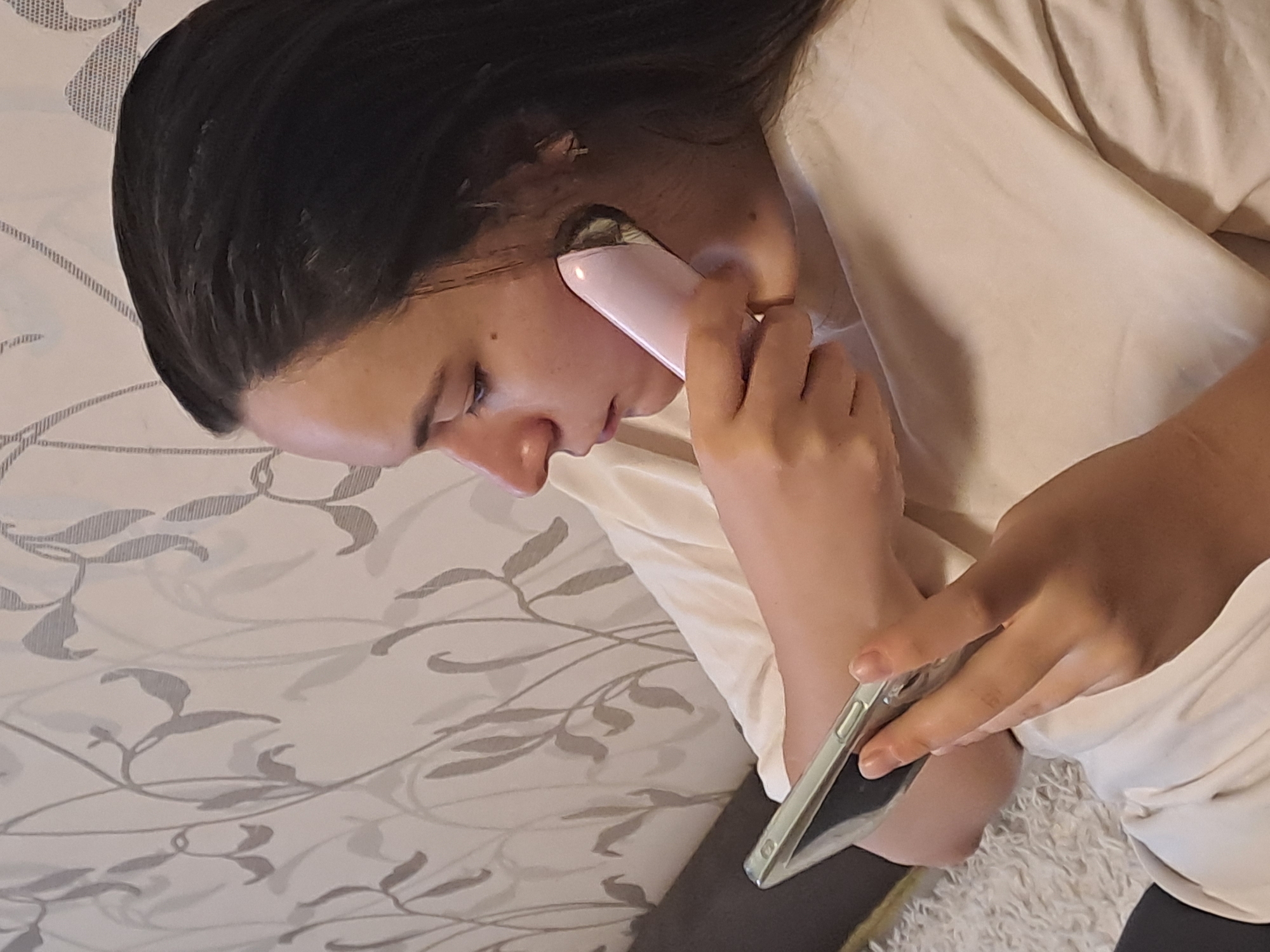 A person is lying on a bed with a patterned bedsheet. The person is holding a pink device to their face with their right hand, near their ear. In their left hand, they hold a smartphone. The person has dark hair and is wearing a light-colored t-shirt. The scene is indoors. There is no indication of this being Elektrostal, Russia.