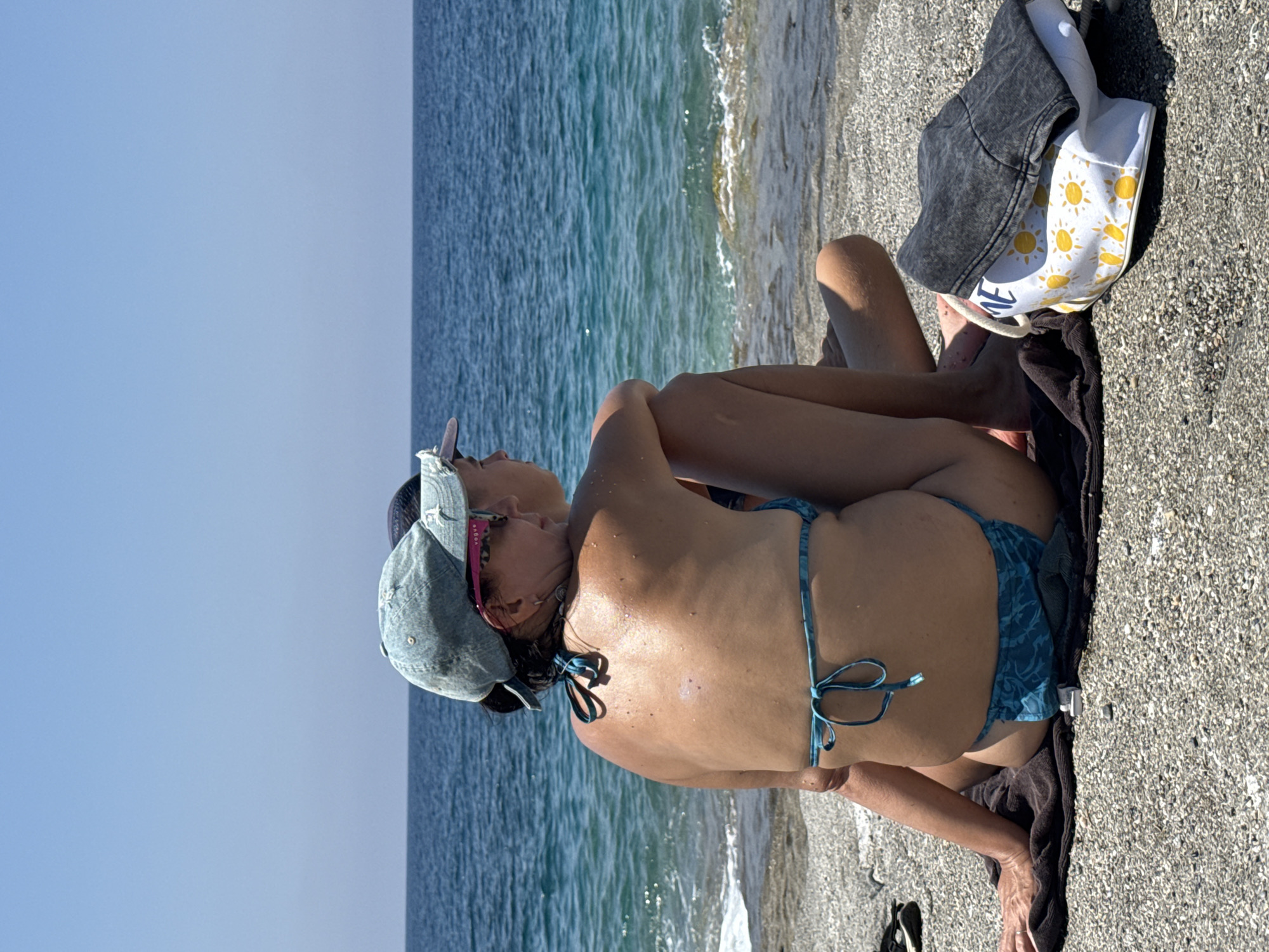 The image depicts two individuals sitting on a pebble beach. The person in the foreground, viewed from the back, is wearing a light blue bikini and a light blue baseball cap. This individual's arms are crossed, and their legs are bent. Beside them, slightly behind, another person is also seated, wearing a dark baseball cap and facing the ocean. Between the two individuals and to the right, a white tote bag with a yellow sun pattern is visible, along with a folded dark gray garment, possibly a jacket or a hat. The background consists of the bright blue sea meeting the clear sky. The scene is illuminated by bright sunlight, casting shadows on the beach. The overall environment is that of a beach with a clear blue sky and calm sea. The exact location in Alanya, Turkey cannot be determined from the visual information provided.