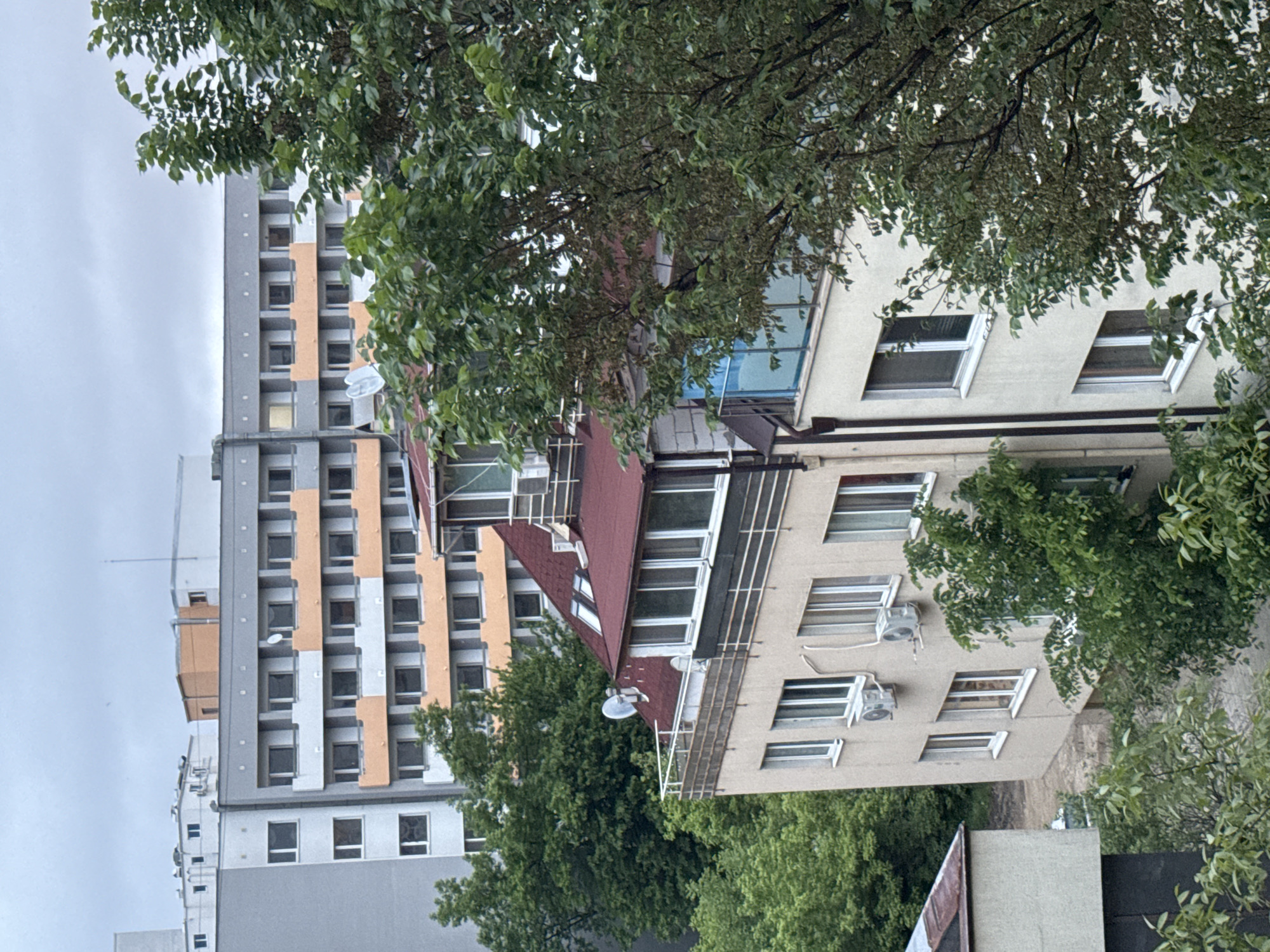 The image depicts a cityscape scene in Sofia, Bulgaria. In the foreground, trees with green foliage obscure part of a beige building with multiple windows and air conditioning units on its exterior walls. The building has a red-tiled roof extension. Adjacent to the beige building is a taller, grey apartment building with alternating grey and orange facade sections and numerous windows. The sky is overcast. No people are visible.