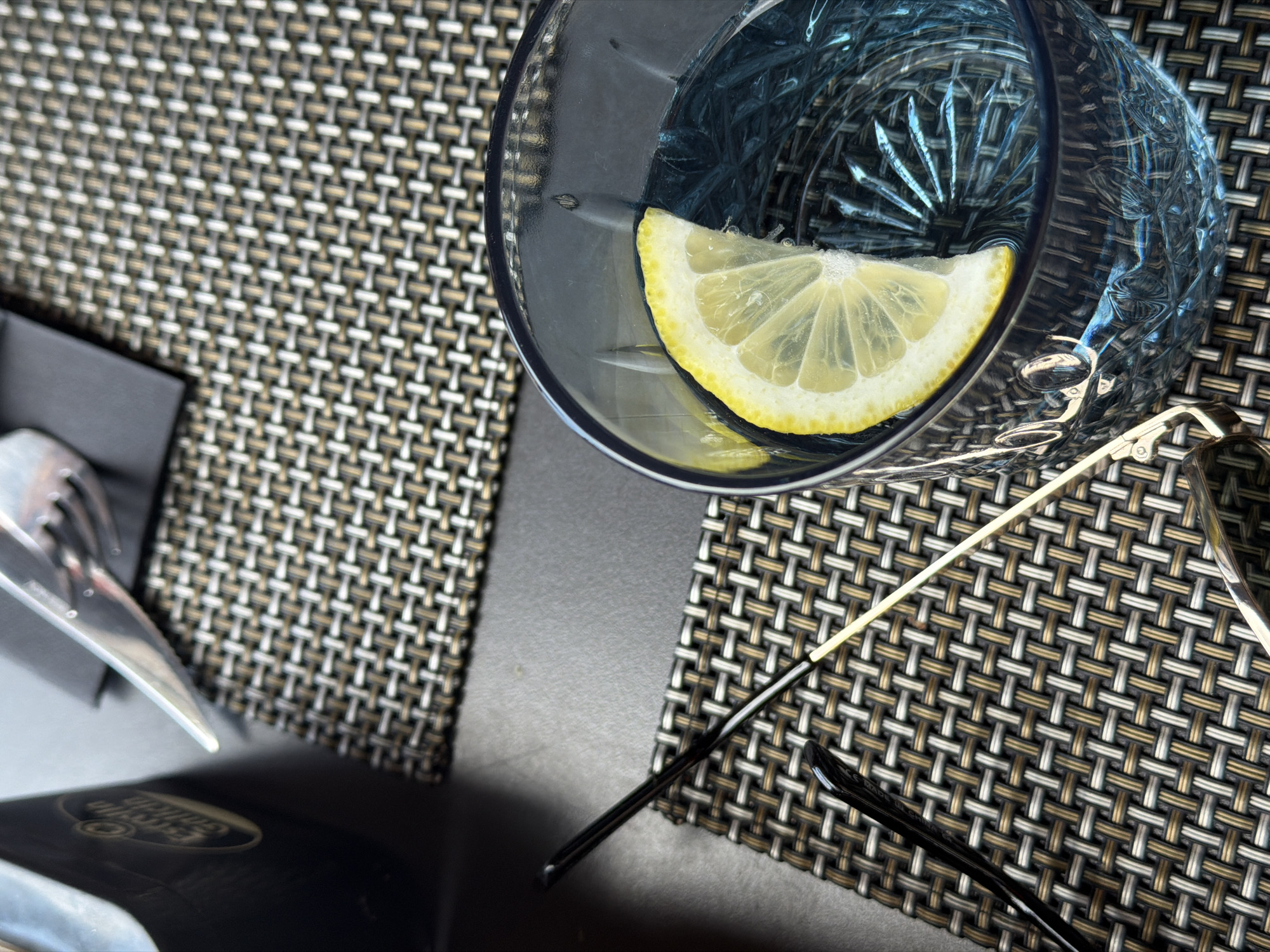 This is a close-up, overhead shot of a table setting with a woven placemat, a glass of water with a lemon slice, and a pair of eyeglasses. The glass is clear with a blue tint and has a faceted design. The lemon slice is bright yellow and positioned in the upper right quadrant of the glass. The eyeglasses have thin, gold-colored arms and dark frames. In the lower left of the image, a silver fork rests on a black napkin. The background is a textured, woven placemat with alternating brown, white, and grey threads. The lighting suggests it could be daytime, possibly indoors or on a shaded patio, given the soft light. The setting appears to be a restaurant or cafe, indicated by the cutlery and placemat. No discernible text is visible in the image.