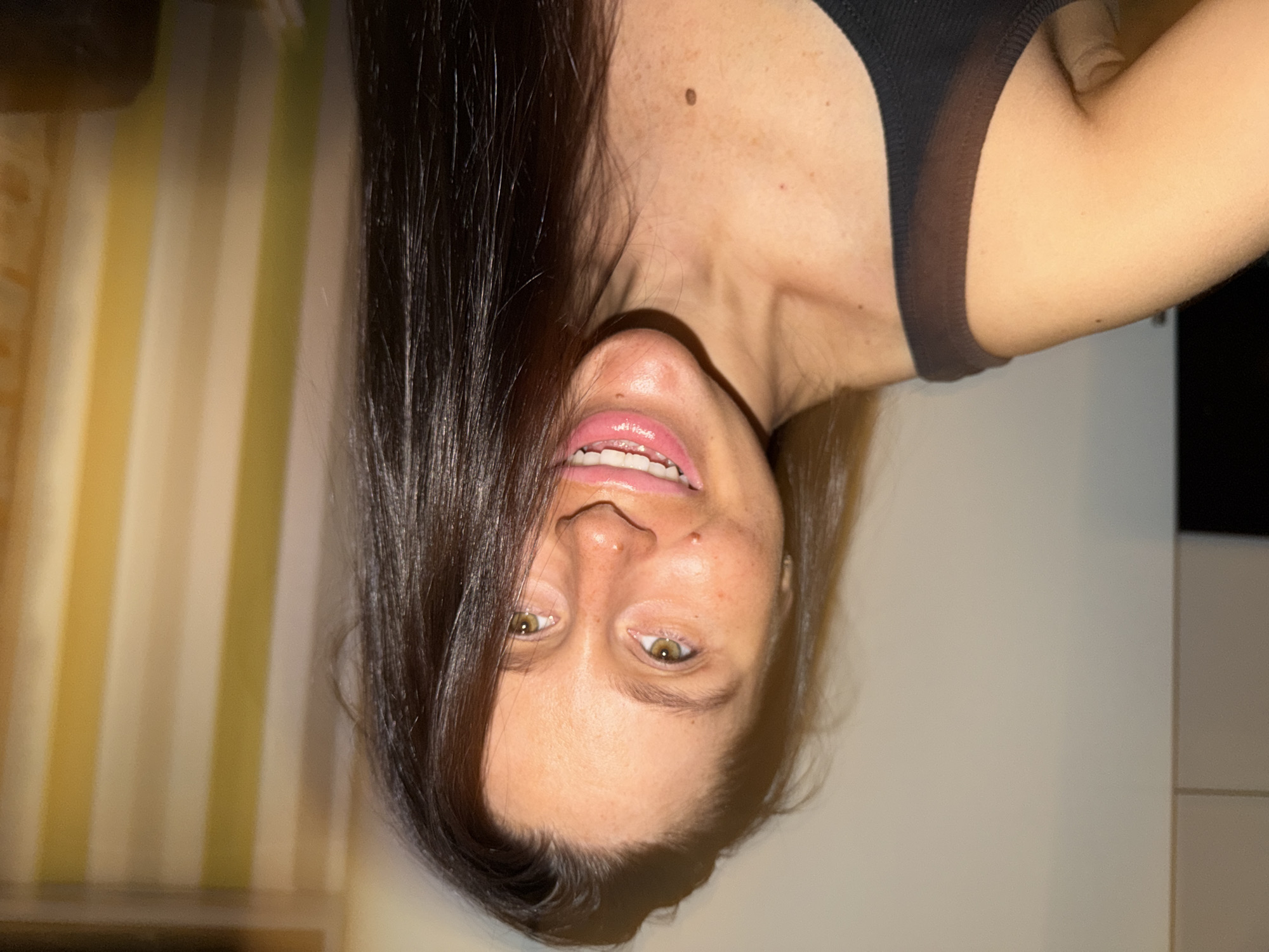 A woman with long dark hair and fair skin is pictured upside down. She is wearing a black top, and her shoulder and arm are visible on the right side of the frame. Her face is centered, displaying a smile and hazel eyes. The background includes vertical stripes of yellow and white on the left, and a plain white wall on the right with a hint of light-colored tiling or paneling at the bottom right corner. There is no indication of the location being Khimki, Russia.