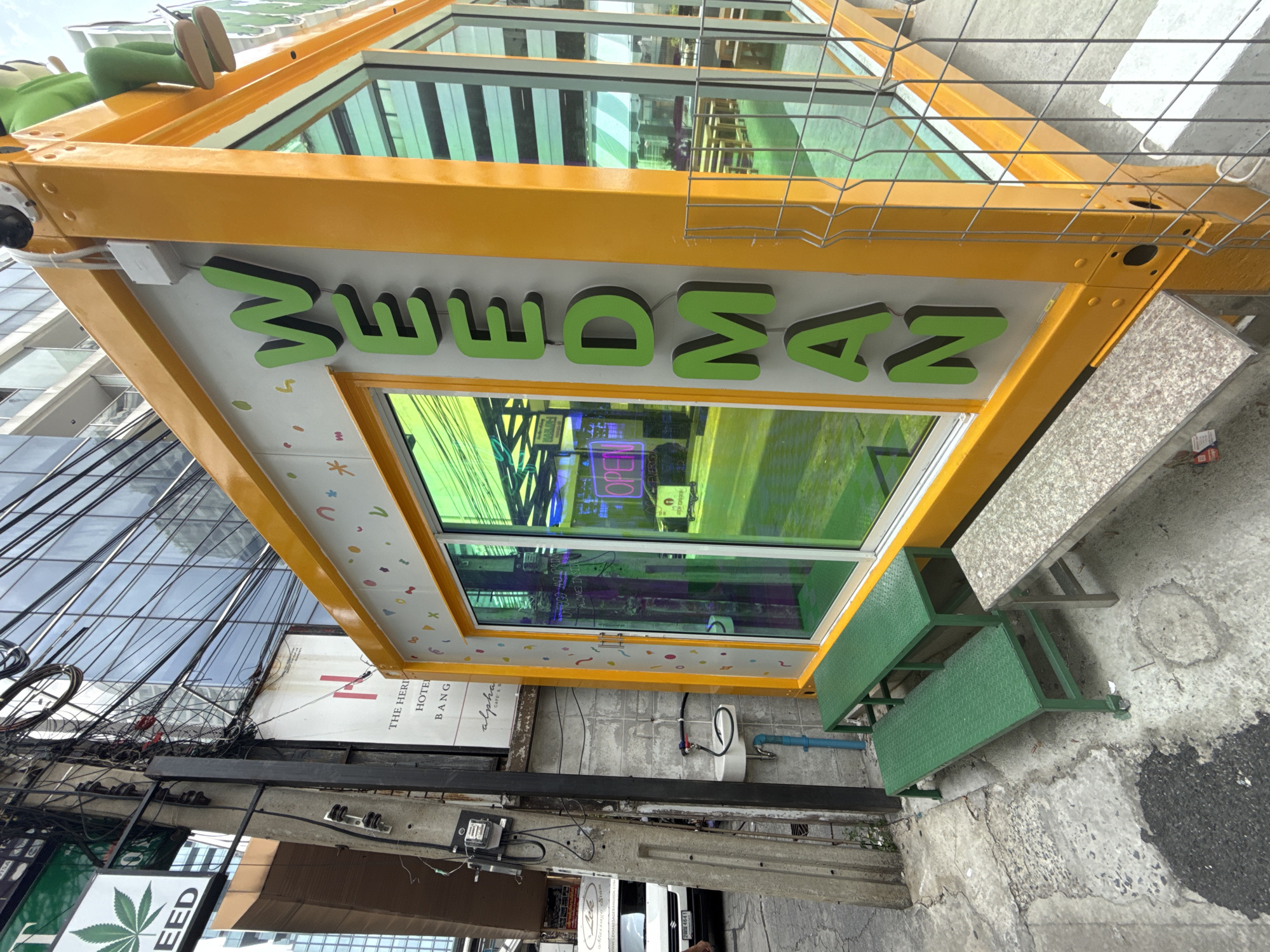 An eye-level outdoor shot captures a vibrant street scene in a dense urban environment, consistent with Bangkok, Thailand. The primary subject is a small, standalone commercial shop characterized by a bright yellow frame and large, green-tinted glass windows.
Above the main window, a prominent sign features three-dimensional green letters. The letters are stylized, with the initial characters resembling upside-down 'W's or stylized 'M's, followed by 'M', 'A', and 'N'. A possible interpretation of the full word is "MAMAWAN" or "WWWMAN". On the very top of the yellow structure, the lower half of a cartoonish figure, with green legs and beige shoes, is visible.
In front of the shop, a two-tiered green metal bench or step is present. The sidewalk is concrete, showing signs of wear and tear.
The background reveals a bustling cityscape. To the left, a tall, modern building with a glass facade reflects the sky and surroundings. A dense and intricate network of black utility wires and cables crisscrosses poles and structures, a common feature in many Southeast Asian cities. Below these wires, several other signs are partially visible: one white sign reads "THE HERITAGE HOTEL BANG...", indicating a nearby hotel, and another sign displays a green cannabis leaf logo with the word "SEED", suggesting a cannabis dispensary, which is legal in Thailand.
Reflections in the shop's tinted windows show parts of the street and an interior neon sign clearly displaying the word "OPEN". The scene is brightly lit, indicating it is daytime with clear weather. The overall impression is one of a lively commercial district with a blend of modern establishments and characteristic urban infrastructure.