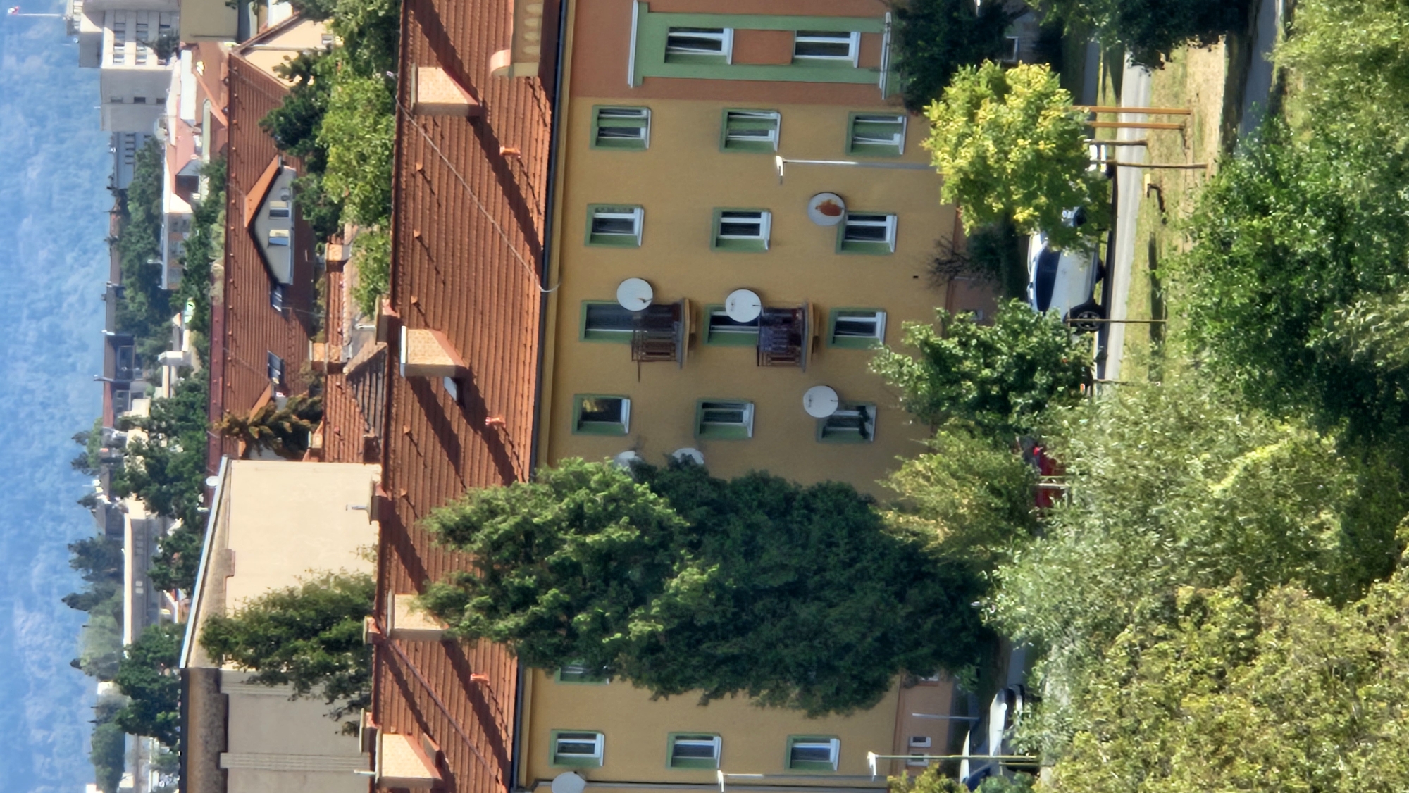 The image shows a residential building in the city of Prešov, Slovakia. The building is painted yellow with red tile roofs and green shutters on the windows. There are satellite dishes on the exterior walls. In front of the building, there is a tall evergreen tree, and several other trees and bushes. In the foreground, there are cars parked on a street, and in the background, there are other buildings and a distant cityscape. The scene appears to be a sunny day.