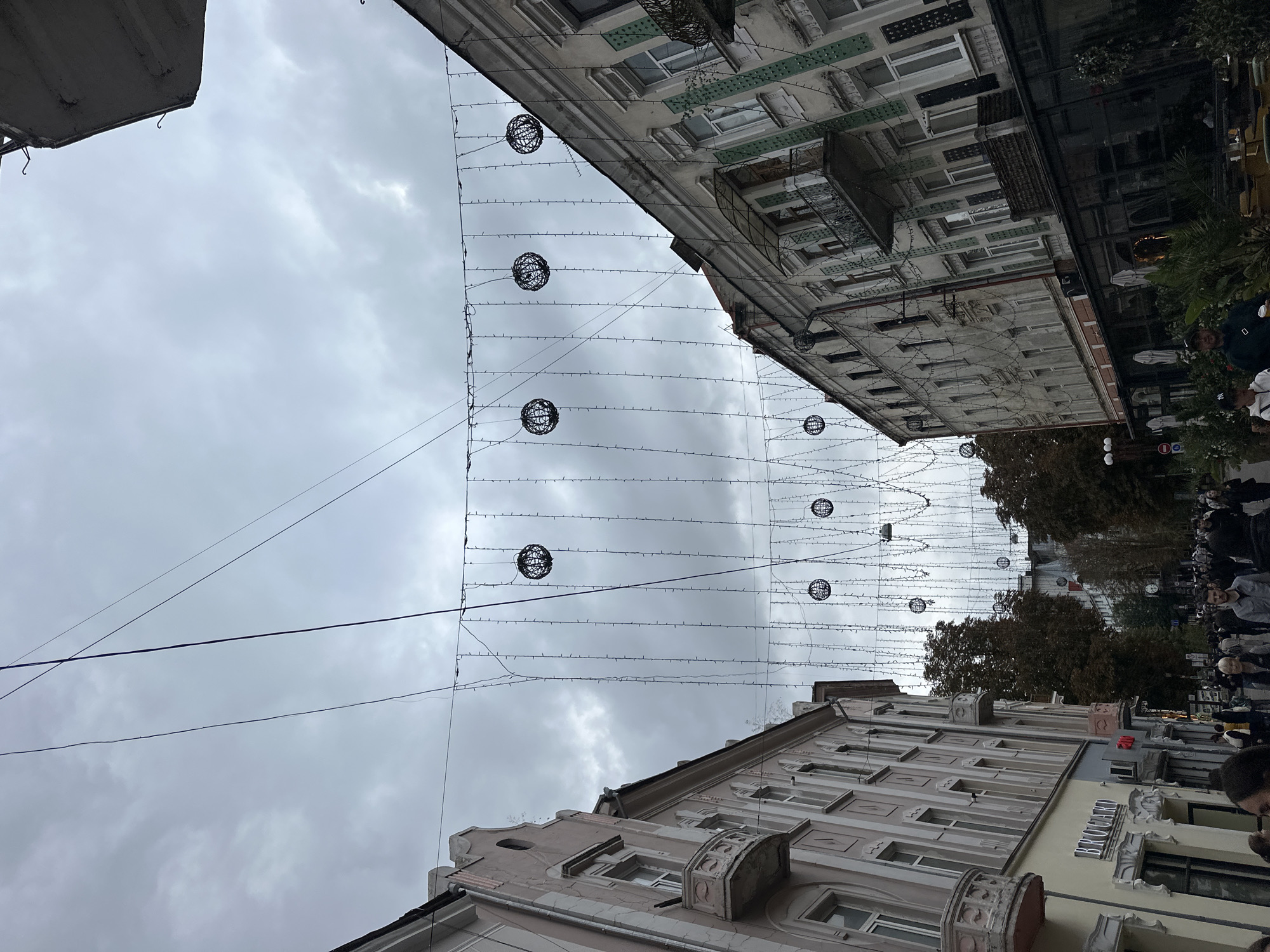 The image depicts a street scene in Ternopil, Ukraine, under a cloudy sky. The foreground shows a street lined with ornate buildings on both sides, featuring balconies and elaborate architectural details. Strings of lights and decorative spherical light fixtures are suspended across the street, suggesting a festive or decorative purpose. Numerous people are walking along the street, some appearing to be shoppers or pedestrians. On the left side, a building has a sign that reads "BROCARI." The overall impression is of a busy urban street, possibly a commercial district, adorned for an event or season.