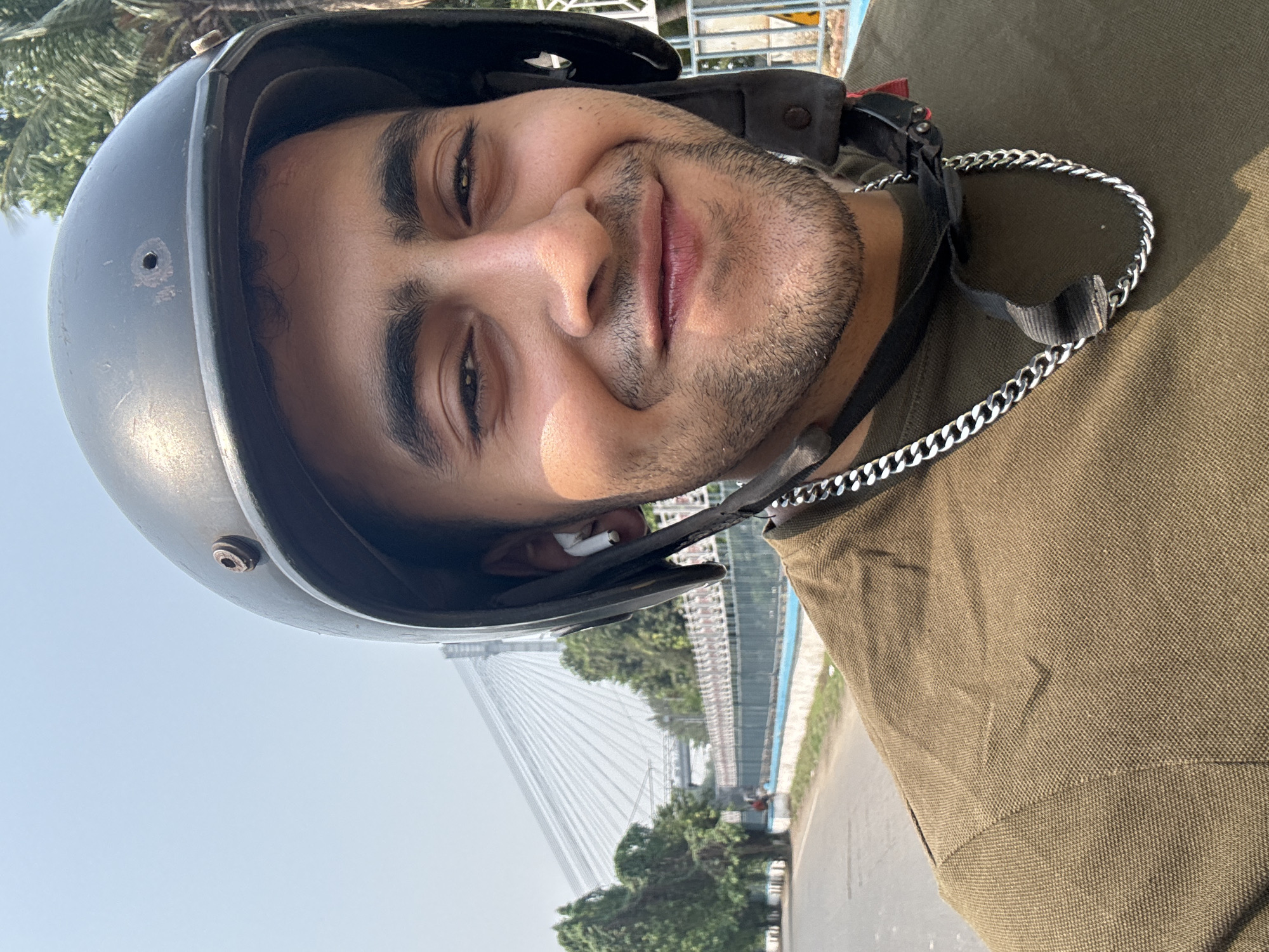 A medium-shot selfie captures a smiling South Asian male wearing a grey helmet and an olive green shirt. He has dark hair, dark eyebrows, and a neatly trimmed beard. A silver chain hangs around his neck. An earbud is visible in his left ear. In the background, the Vidyasagar Setu bridge, a landmark in Kolkata, India, is partially visible, suggesting that the photo was taken in close proximity to it. Trees line the road. The sky is overcast.