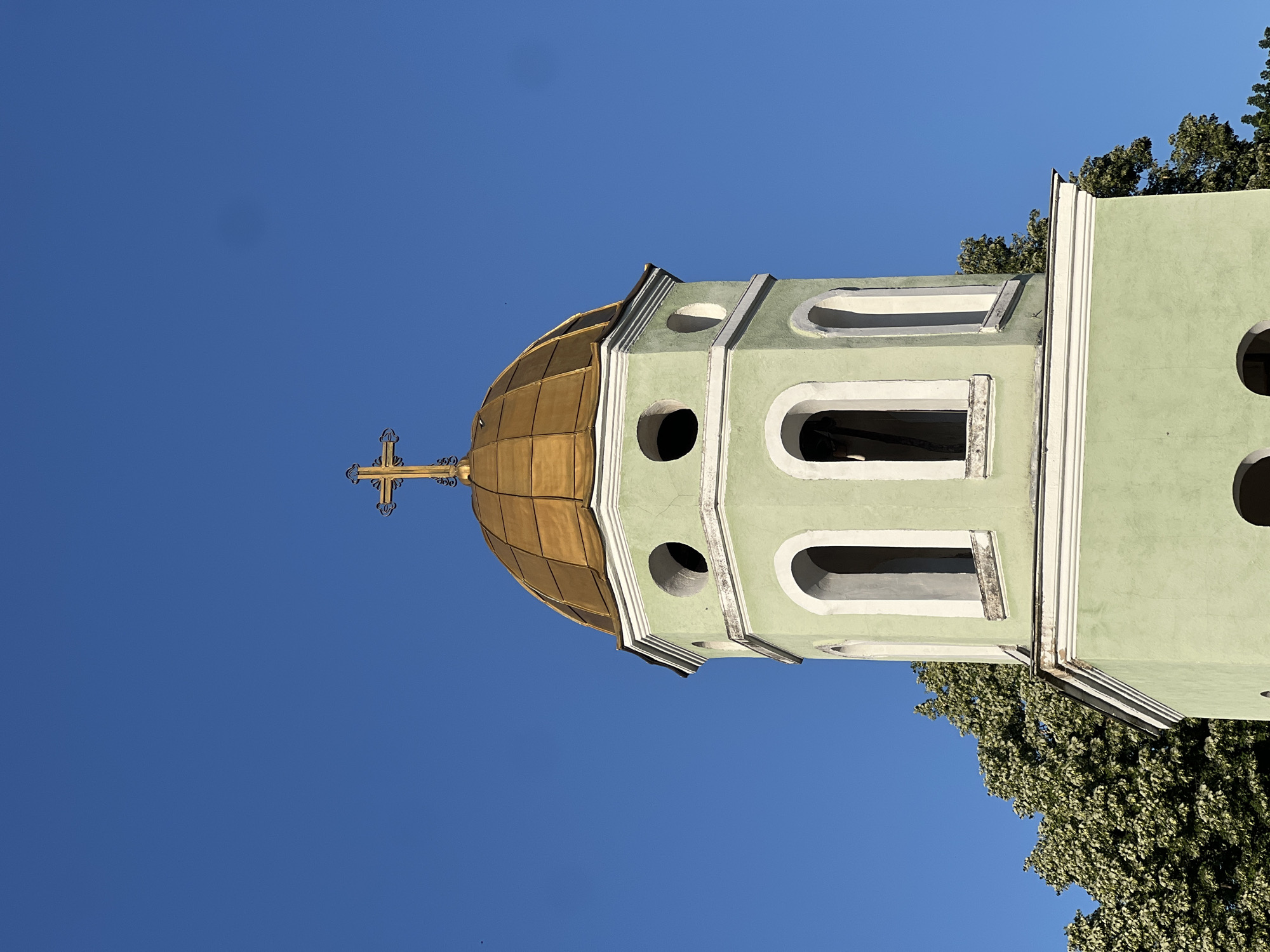 A close-up, vertically oriented shot focuses on the top of a green bell tower topped with a golden dome, against a clear, bright blue sky. The bell tower has a light green facade with white trim accentuating arched windows and circular openings. A ornate, golden Orthodox cross crowns the dome. To the right of the bell tower, lush green foliage from a tree reaches into the frame. The bright sunlight suggests it is daytime, likely mid-morning or afternoon. The setting appears to be Zelenikovo, Bulgaria, hinted by the architectural style and the presence of Orthodox Christian symbolism. There is no visible text in the image.