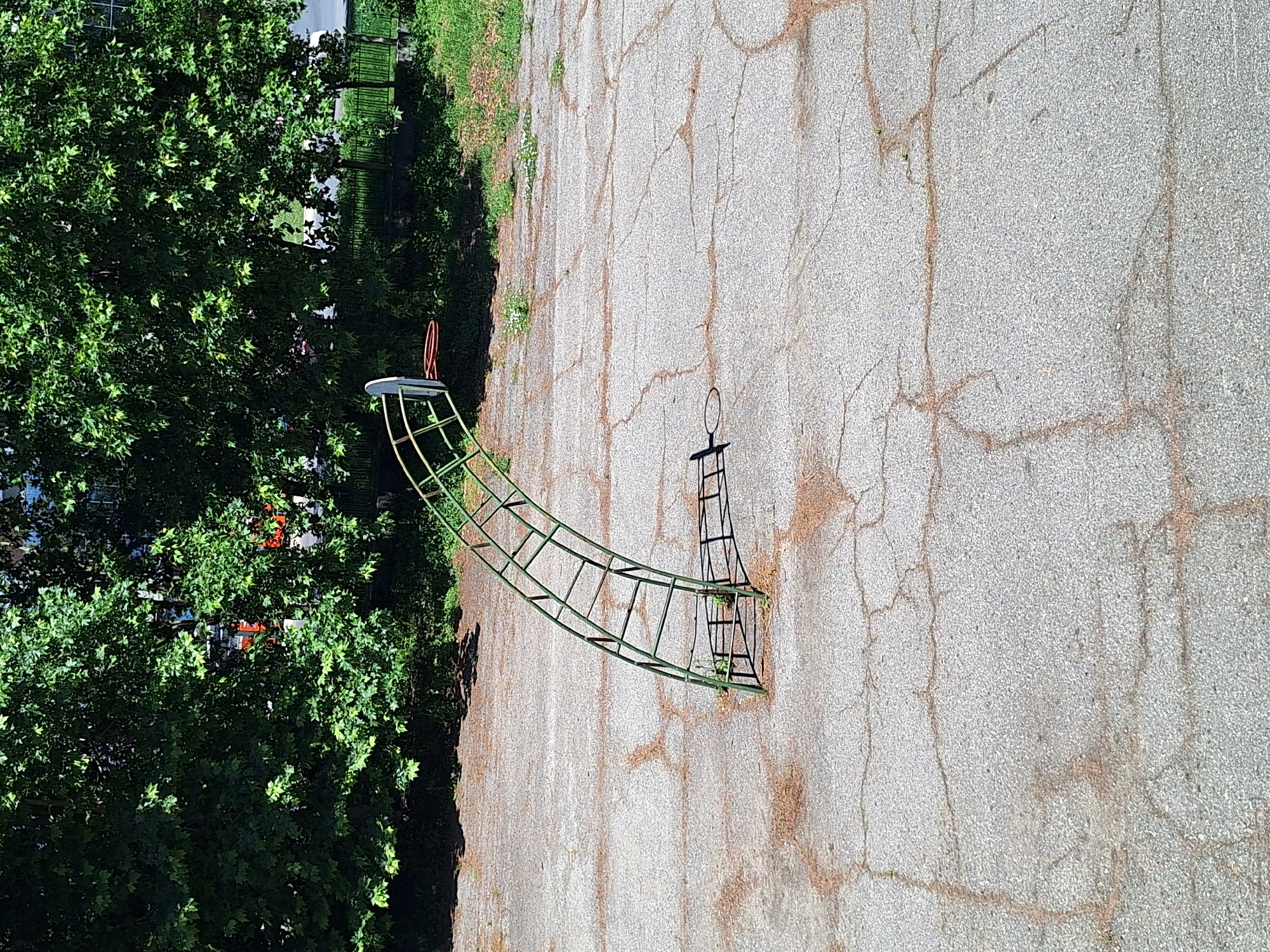The image shows a playground structure, possibly a climbing frame or a small slide, in an outdoor setting. The structure is made of green metal bars and has a curved, ladder-like section leading to a flat platform. A red hoop is attached to one end of the structure. To the right of the structure, there is a black metal frame with a hoop on top, resembling a mini basketball hoop or a decorative element. The playground is situated on a cracked asphalt surface, with dry leaves and debris scattered around. To the left of the asphalt, there is a lush green tree with dense foliage, casting shadows on the ground. In the background, through the leaves of the tree, glimpses of buildings with orange and white elements can be seen. A metal fence and some grass are also visible behind the tree. The lighting suggests it is daytime, likely sunny, with shadows indicating the sun is at an angle. The scene appears to be in a park or a residential area in Varna, Bulgaria. There are no people visible in the image, and the playground equipment seems to be unused at the moment. The overall impression is one of a quiet, possibly late afternoon or early morning scene in a public space.