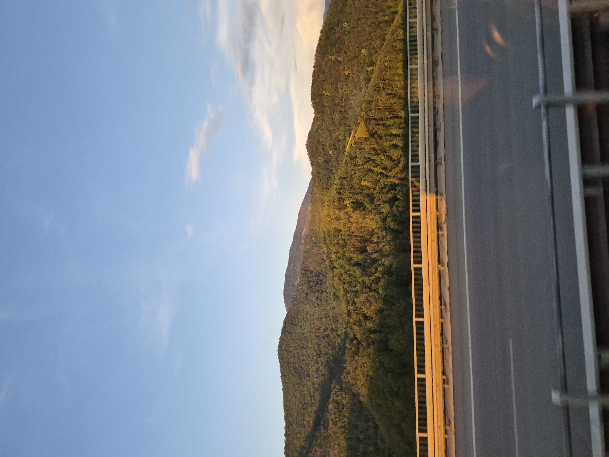 A scenic view captures rolling, verdant hills bathed in the warm glow of sunset, under a clear blue sky with a few wispy clouds. In the foreground, a modern highway stretches across a bridge, complete with a guardrail and distinct lane markings. The scene evokes a sense of peaceful travel and natural beauty, with the golden hour light highlighting the textures of the forest-covered slopes. The location is the vicinity of Vrachesh, Bulgaria.